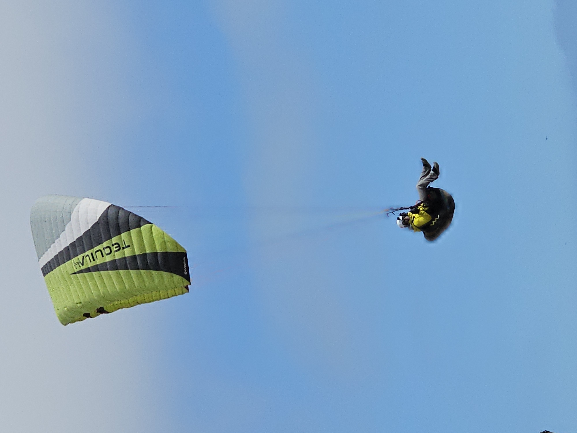 The image shows a paraglider in the sky in Alto Hospicio, Chile. The paraglider is upside down with their feet in the air. The paraglider's wing is partially collapsed.