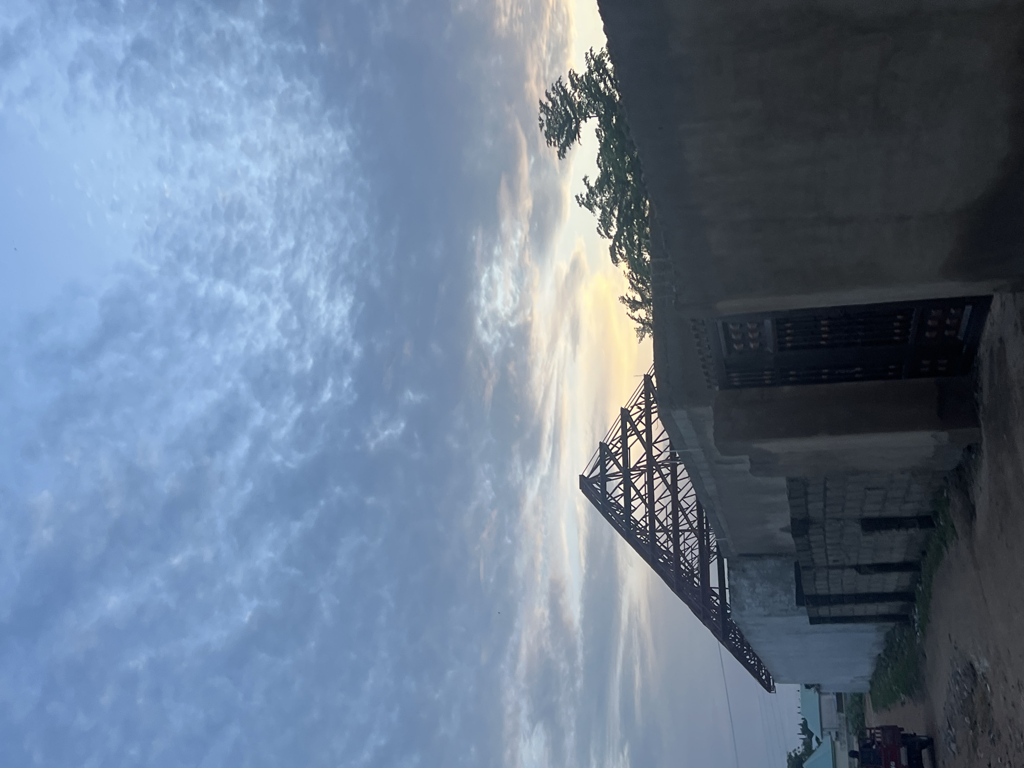 The image depicts a building under construction in Jalingo, Nigeria, as indicated by the presence of exposed roof trusses. The building features concrete walls and a dark metal gate. In the foreground, a dirt road is visible, with a small red truck carrying a person in the rear on the left side of the frame. The sky is filled with textured clouds, suggesting either dawn or dusk, with a hint of yellow and orange light peeking through the clouds on the horizon. Trees are visible behind the wall, and other buildings with light blue roofs can be seen in the distance. The overall scene is one of a developing urban area.