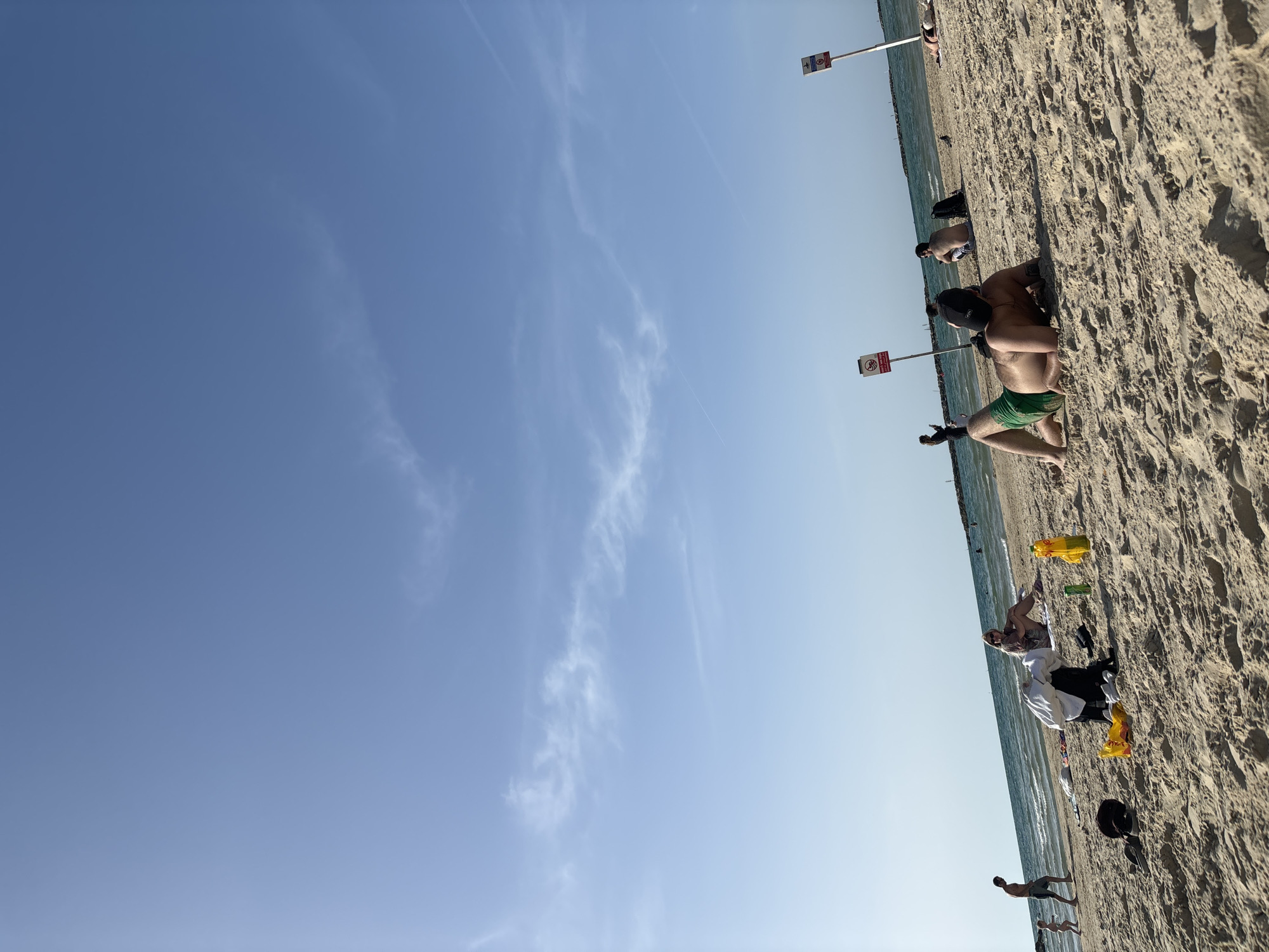 The image captures a sunny day at a beach in Tel Aviv, Israel. The scene includes several people scattered along the shoreline. Individuals are seen engaging in typical beach activities such as sunbathing, standing, and walking. Beach accessories like clothing items, bags, and water bottles are strewn on the sand. Warning signs are fixed along the beach. The sky is clear, with sparse wisps of clouds.