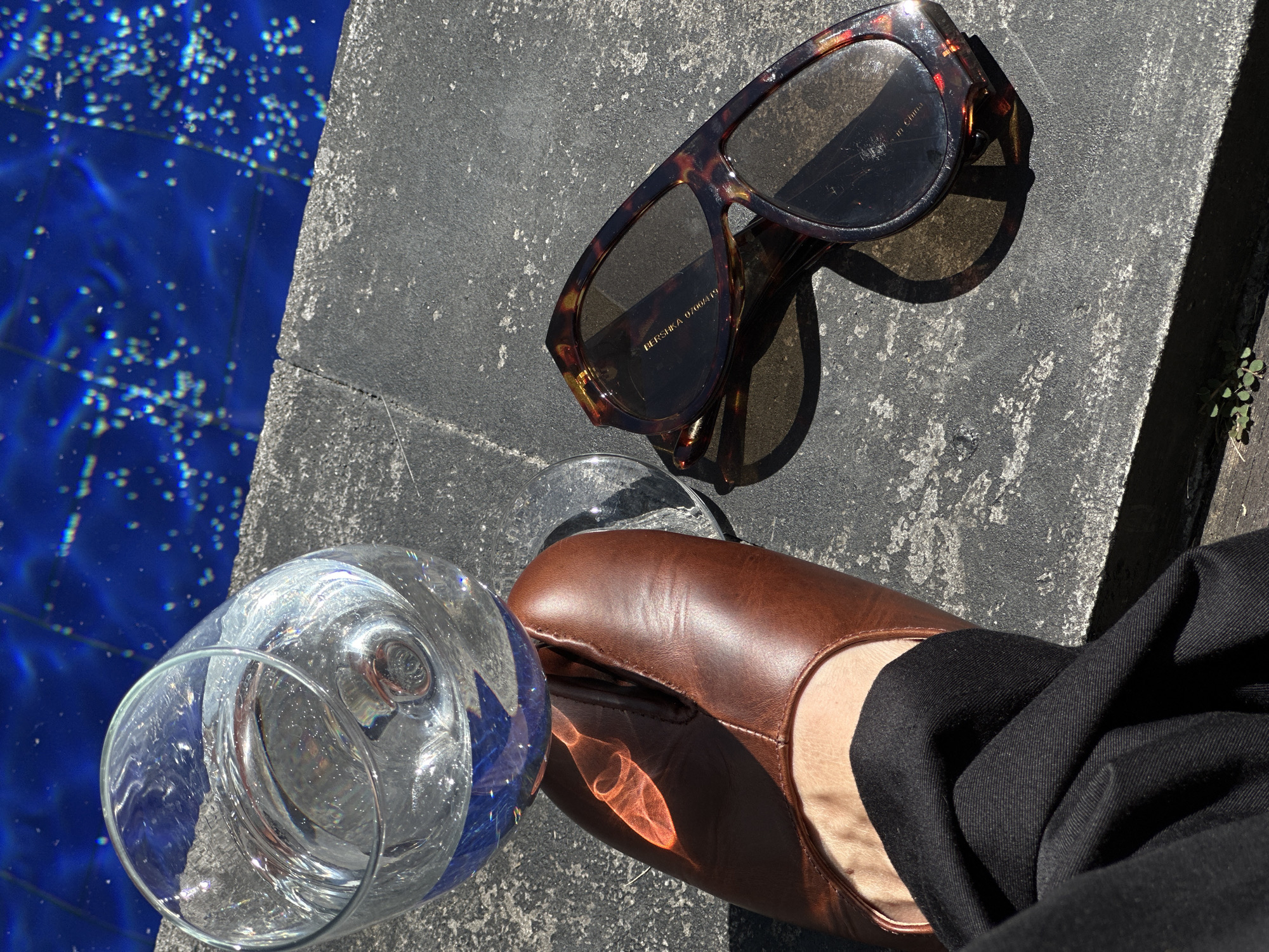 A close-up, overhead shot shows a person's left foot wearing a brown leather split-toe shoe. The shoe rests on a dark grey, textured surface next to a glass of clear liquid and a pair of tortoiseshell sunglasses. The background is a bright blue, rippling surface, likely a swimming pool. The scene is illuminated by direct sunlight, casting shadows and creating highlights on the glass and shoe. The location is identified as North Kuta, Indonesia.