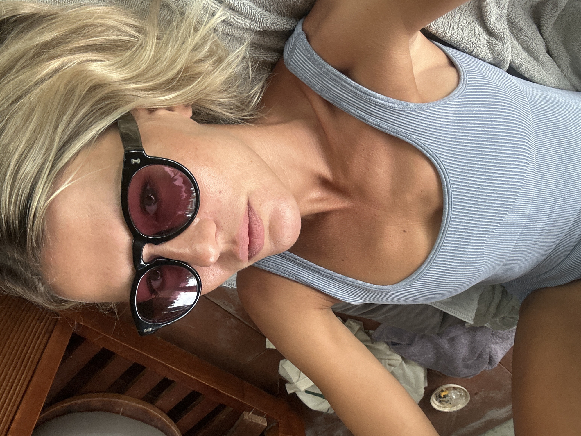 A woman with blonde hair lies on her back, looking directly at the camera through black sunglasses with dark red-tinted lenses. She wears a light blue and white striped ribbed tank top, and her tanned skin is visible. Her head rests against a grey, textured fabric, which also appears around her shoulders. To the left and below her right arm, a wooden surface is visible, possibly part of the floor or furniture. In the lower right corner, on the wooden surface, there is a small, clear, round dish containing some residue. Below her, crumpled grey and purple textiles are scattered on the wooden floor. The reflections in her sunglasses suggest an indoor setting, possibly showing a ceiling fixture. The wooden elements in the scene evoke a natural or traditional architectural style. The overall setting appears relaxed and informal. No specific activity is taking place beyond posing for the photograph. The location is Mengwi, Indonesia. There is no visible text.