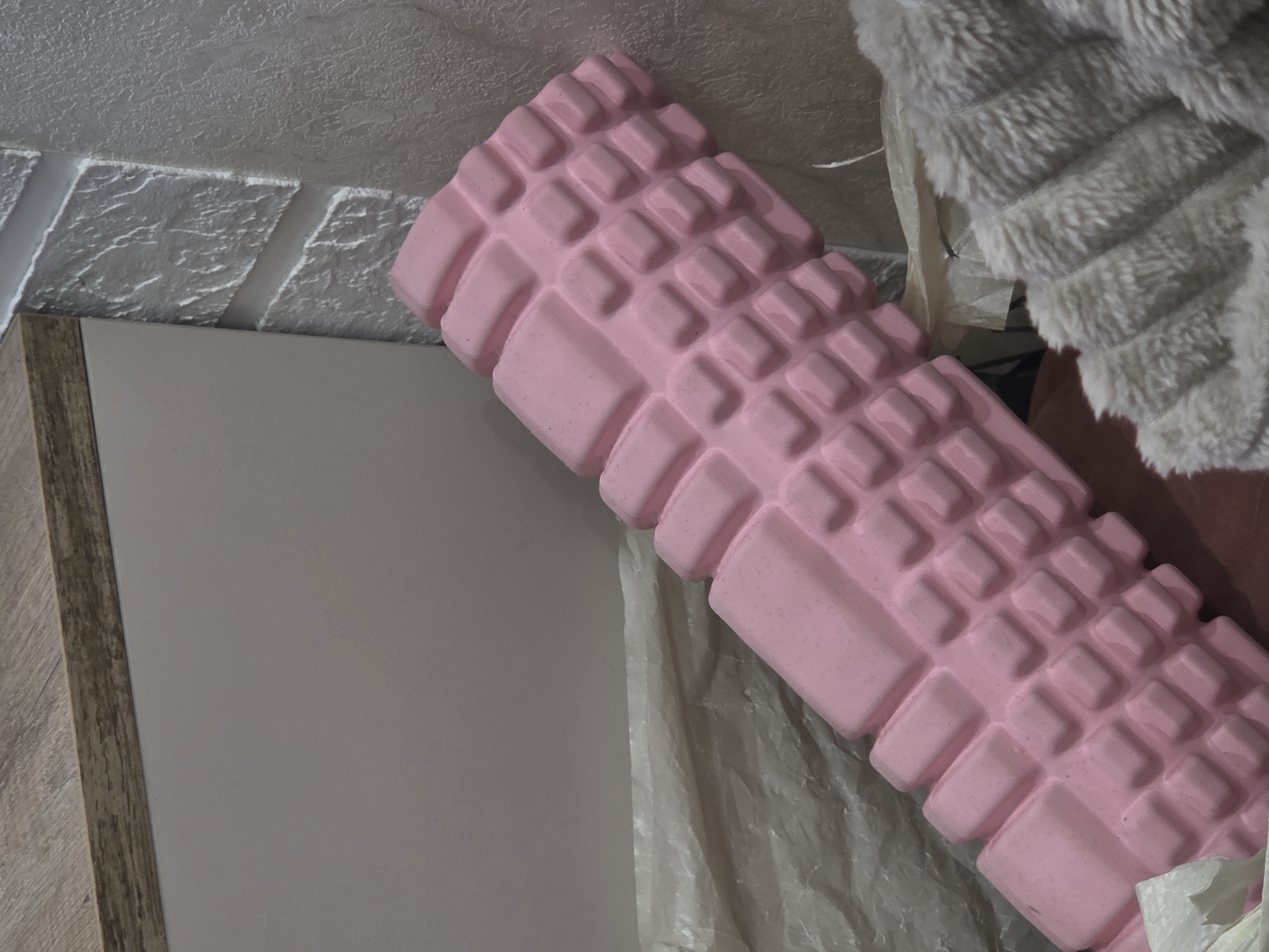 The image shows a pink foam roller with a grid pattern on a surface that includes a white wall with decorative brick-like indentations, a cream-colored surface with a wood-grain frame, crumpled white tissue paper, and a light gray fuzzy fabric. The environment appears to be indoors.