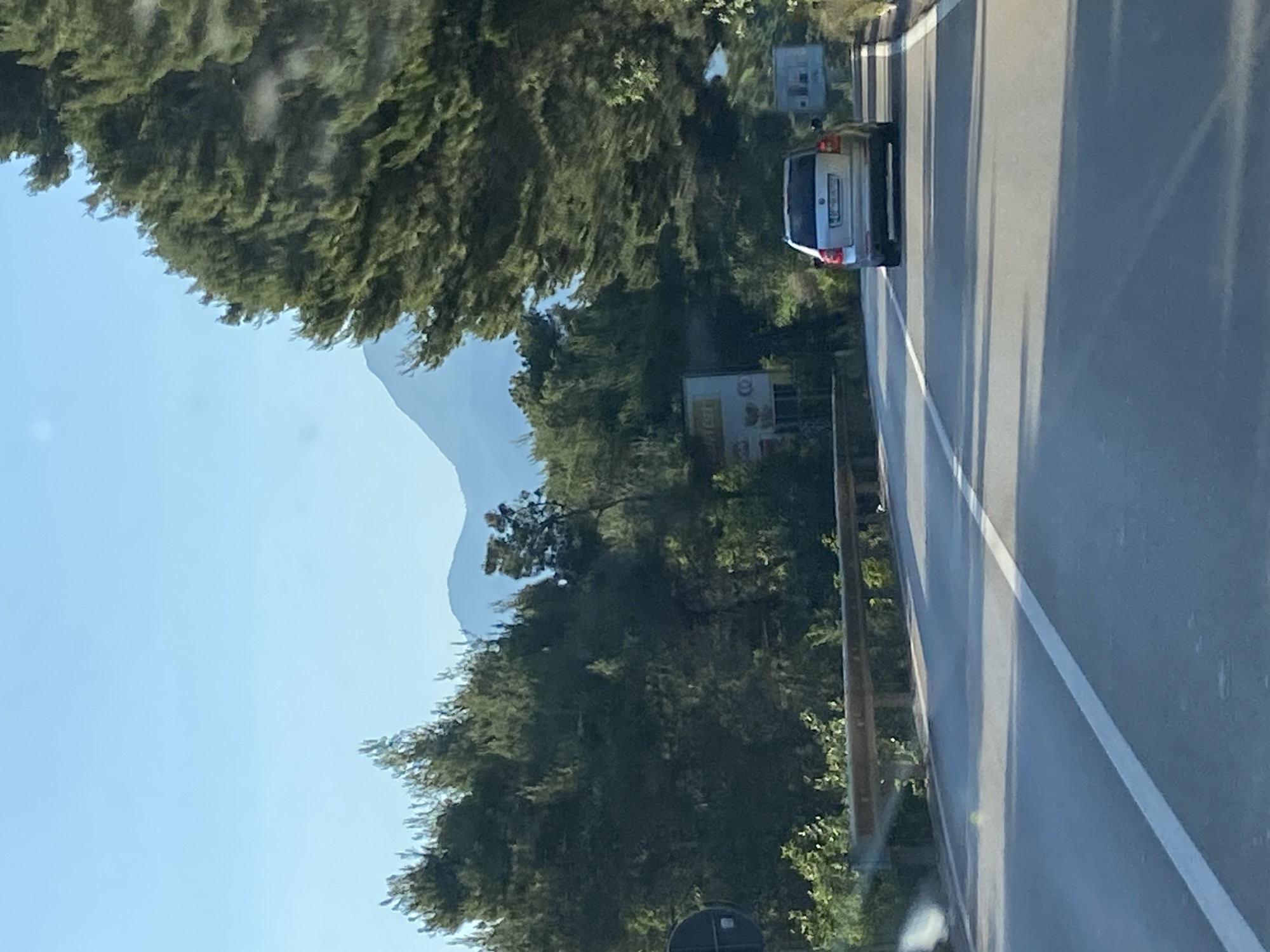 The image depicts a roadside scene near Blizikuće, Montenegro. A silver car is traveling on a paved road with a solid white lane marking. The road is bordered by lush green trees and vegetation. In the background, a distant mountain range is visible under a clear blue sky. A roadside sign is also present. The image appears to have been taken from within another vehicle.