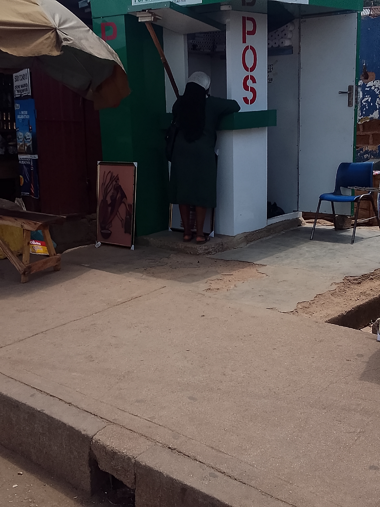 A woman wearing a dark green dress and a white head covering is at a Point of Sale (POS) station located on a sidewalk in Bukuru, Nigeria. The POS station is green and white with the letters "POS" in red on the side. The woman is leaning against the counter, interacting with the device. To the left of the station, there is an artwork leaning against the wall. A blue chair stands to the right of the POS station. A small wooden table and an umbrella are located further to the left. The sidewalk is concrete. In the background, there are signs visible in a shop.