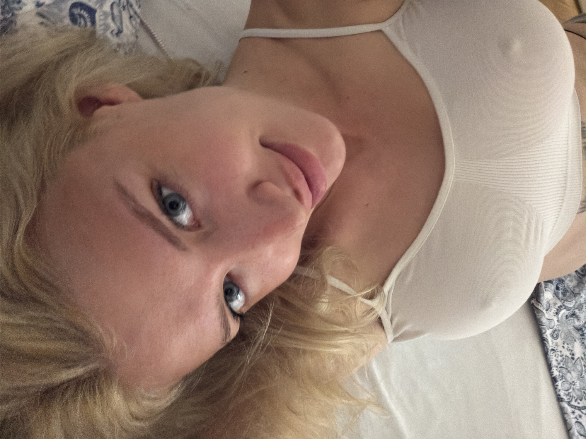 A blonde-haired woman with blue eyes lies on her back in what appears to be a bed, wearing a white tank top. The bed has white sheets and a blue and white patterned pillow or blanket is visible. The photo is taken from a close, overhead perspective, focusing on the woman's face and upper body. There is no indication of the location being in Belgrade, Serbia.