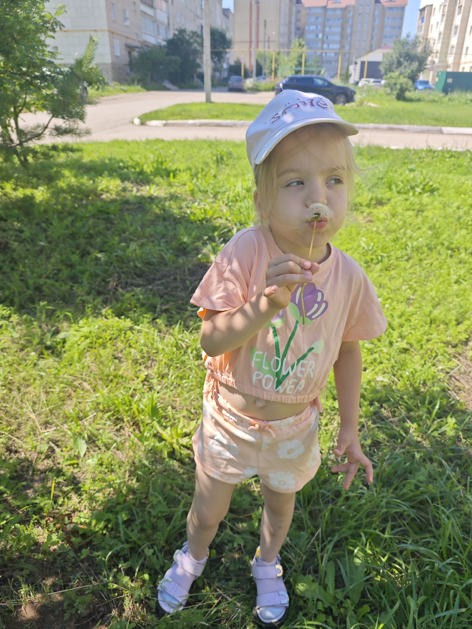 A young Caucasian girl stands in a grassy area, blowing on a dandelion. She is wearing a pink t-shirt with a flower graphic and the words "FLOWER POWER," matching peach shorts with a floral pattern, and light purple sandals. A white baseball cap is on her head. In the background, residential buildings and a road are visible, suggesting an urban environment. The scene is consistent with a park or residential courtyard in Tujmazy, Russia, on a sunny day.