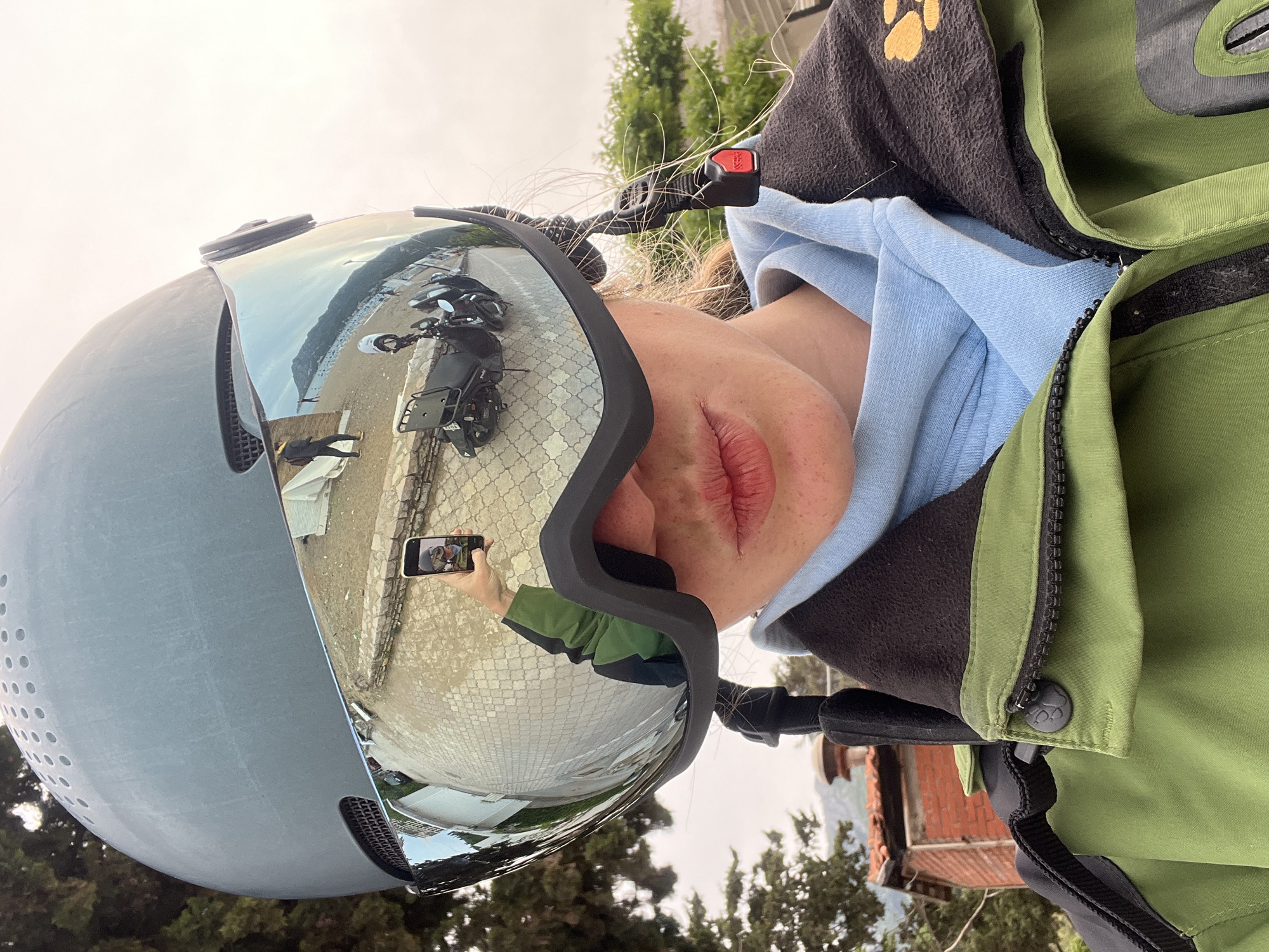A person wearing a helmet with reflective goggles is shown. The goggles reflect a street scene with a motorcycle parked on a paved area adjacent to a beach. The person is also wearing a hooded sweatshirt and a green jacket. The location is possibly a coastal city with trees and buildings visible in the background.