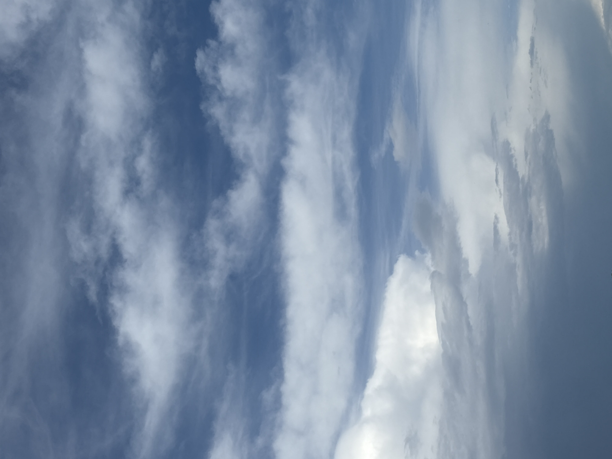 The image shows a daytime sky in Chaikovsky, Russia, featuring a mix of cirrus and cumulus clouds. The sky is predominantly blue, with streaks of cirrus clouds thinly spread across the upper portion. Thicker cumulus clouds occupy the right side of the image, some appearing brighter white, and others darker grey, suggesting varying levels of density and potential precipitation. There are no people or objects visible besides the clouds and the sky. The environment is an open-air scene.