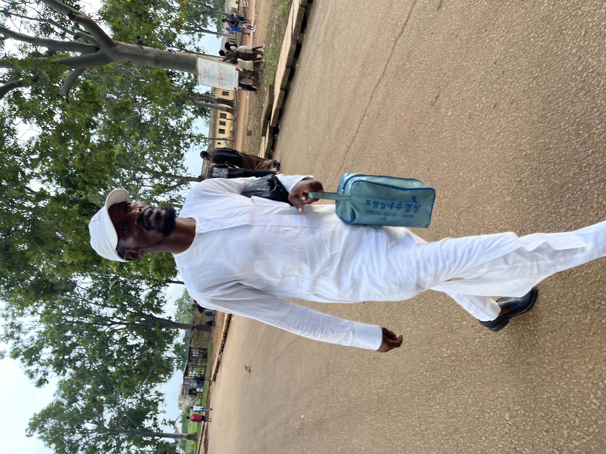 An adult man walks along an unpaved, light brown path or road, flanked by large, leafy trees. He is dressed in a flowing white long-sleeved tunic and matching trousers, paired with black shoes and a white baseball cap. A black bag is slung over his left shoulder, and he carries a small, light blue bag in his right hand. The blue bag features Korean text, which translates to "Pohang Indoor Swimming Pool" (포항실내수영장), along with a phone number, "0274-9019-0028". The word "TORI" is partially visible on his white cap.
In the background, on the right side of the path, several other individuals are visible, some walking and others standing near a low-rise, light-colored building. A white sign with red markings is also present in the middle distance. The scene is outdoors, under what appears to be an overcast or diffused daytime sky, as indicated by the soft lighting and lack of harsh shadows. The overall setting suggests a semi-urban or campus environment, consistent with the provided location context of Rafin Par, Nigeria. The man's expression is neutral as he walks.