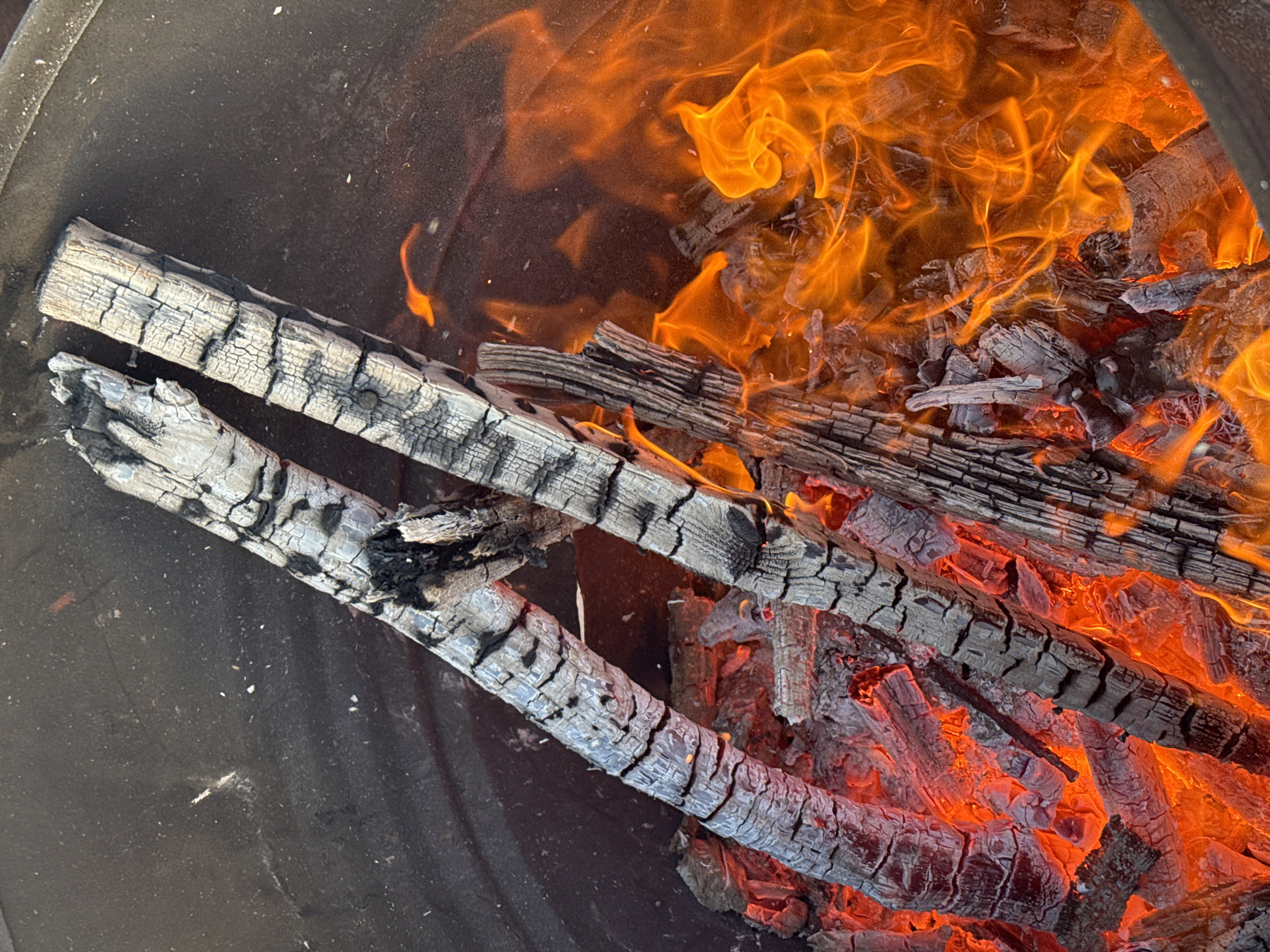 A close-up, high-angle shot depicts burning logs inside a dark metal fire pit or container. The wood is charred, cracked, and ashy, with flames visible both above and below the logs. The bright orange flames contrast with the dark wood and metal. The scene appears contained within a domestic or outdoor setting. Location is not confirmed to be Bol'shoye Kozino, Russia.