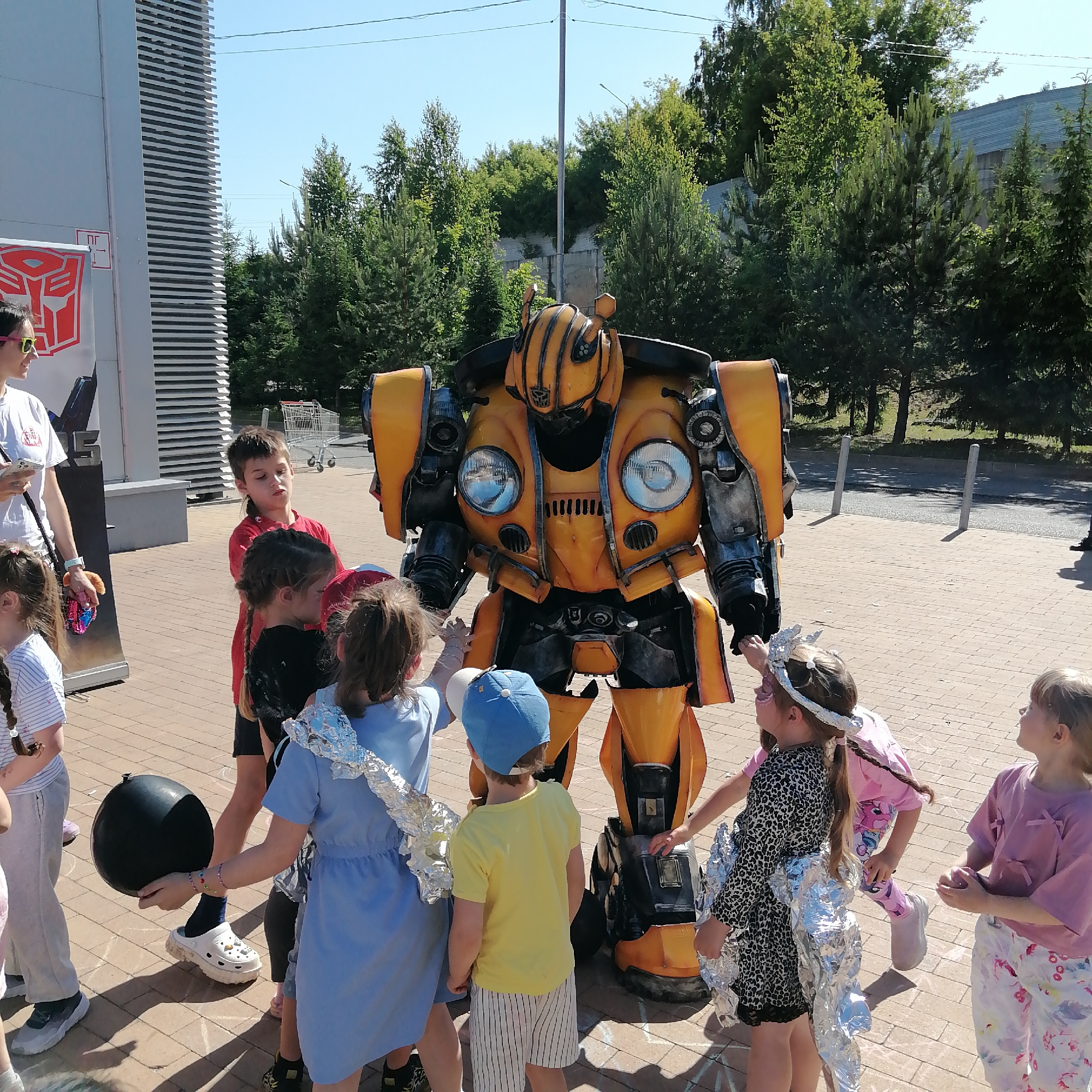 The image depicts an outdoor event in Трубачеевка, Russia, under clear, sunny skies. A person in a detailed yellow and dark gray Bumblebee costume, featuring prominent circular headlights on the chest and the Autobot logo, stands centrally, surrounded by several children. An adult female in a white t-shirt, wearing yellow-rimmed sunglasses on her head, is positioned on the left, observing the scene while holding a smartphone.
The children, of various ages, are actively engaging with the Bumblebee character. Some children are partially adorned with silver foil, suggesting a themed activity or costume element. One girl has pink face paint, and another in a blue dress holds a black spherical object. The ground is a light brown paved surface with visible chalk markings.
In the background to the left, a modern building with light gray vertical slatted panels features a large red Transformers Autobot logo and the Cyrillic text "ПГ" on a white banner. Further back, a line of dense green coniferous and deciduous trees separates the foreground from a visible section of an asphalt road with two light-colored bollards and a distant building with a gray roof. A stack of metal shopping carts is also visible behind the children near the building.