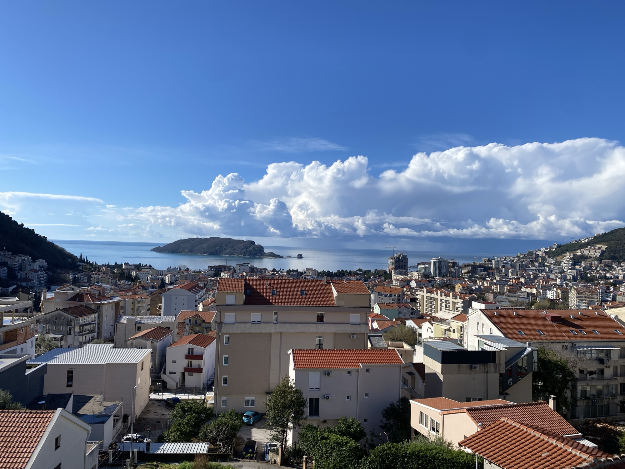 An elevated panoramic view displays the city of Budva, Montenegro, under a clear blue sky adorned with large, bright white cumulus clouds. The foreground is dominated by a dense arrangement of residential buildings, primarily featuring light-colored facades (beige, white, grey) and distinctive red terracotta tiled roofs. Several multi-story structures are visible, some exhibiting modern architectural elements or appearing to be under construction. A light blue compact car and other vehicles are parked in an open lot behind a building in the middle-left. The urban landscape extends towards the calm, light blue waters of the Adriatic Sea. In the mid-distance, Sveti Nikola Island, a large, green, uninhabited island, is prominently situated within the bay, accompanied by a smaller rocky islet to its right. The city's development continues along the coastline and climbs the slopes of green, tree-covered hills that frame the bay on both sides. No individuals are visible.