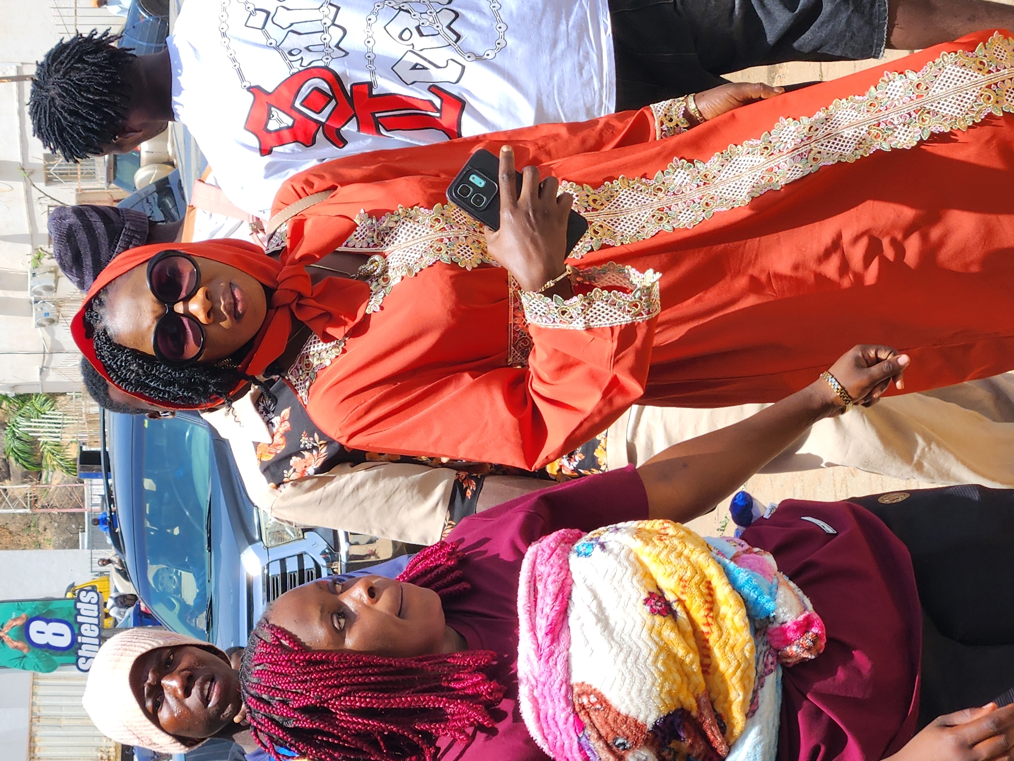 A medium shot captures a group of individuals in an urban environment. A woman, centrally positioned, is clad in an orange dress adorned with intricate lacework. She holds a smartphone in her hand. Nearby, another woman wears a maroon shirt and a wrap featuring vibrant colors. An individual with braids stands closely, and other people are visible in the background. A vehicle is parked in the vicinity. The overall setting suggests an outdoor gathering or public space within a city. A sign for 'Shields' is partially visible.