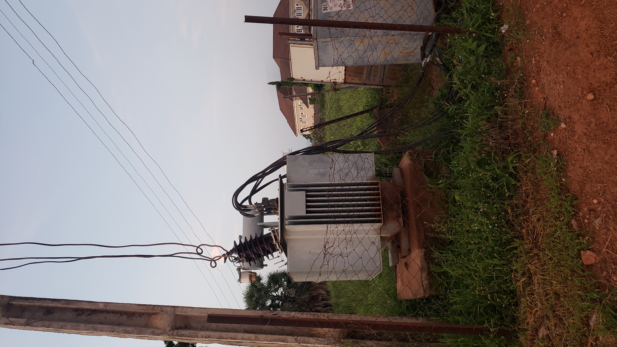The image shows an outdoor scene in Jalingo, Nigeria, featuring an electrical transformer positioned on a concrete base, surrounded by overgrown grass and a chain-link fence. A concrete pole stands to the left, with overhead electrical wires extending across the sky. The transformer is gray and rectangular with ventilation slits on the front. Thick black cables are connected to the top of the transformer and run towards the right. In the background, two buildings with brown roofs and light-colored walls are visible, along with palm trees. Another metallic structure, possibly another transformer or electrical enclosure, is located to the right of the main transformer. The ground in the foreground is a reddish-brown dirt path. The sky is a pale blue, suggesting either early morning or late afternoon light. No people are present in the image.