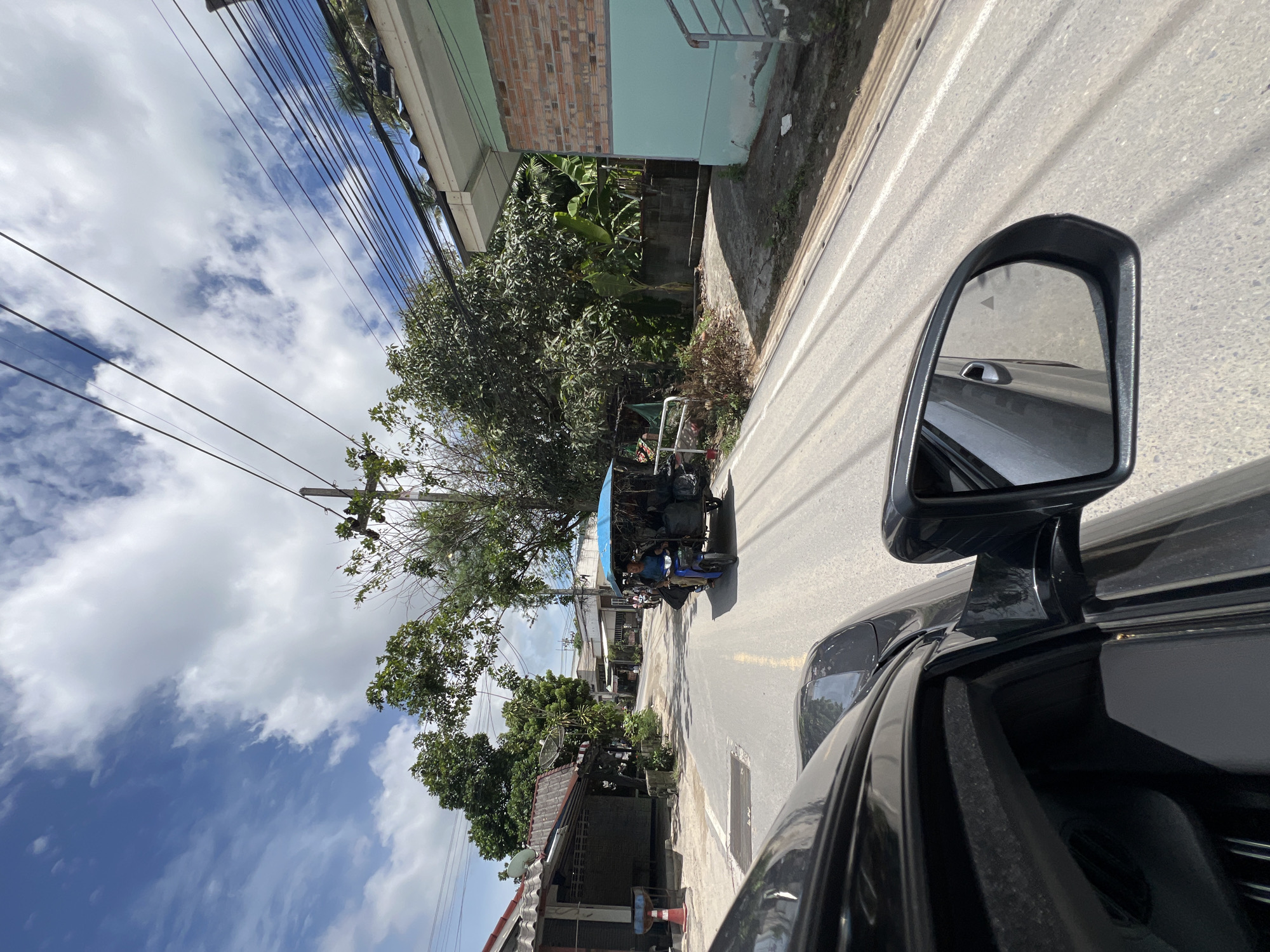 This image, captured from inside a moving vehicle, depicts a street scene in Ban Bang Rak, Thailand. A black car's side mirror and part of its hood are visible in the foreground, reflecting the road and sky. The street itself is paved and appears to be a two-lane road with a yellow dividing line. In the middle ground, a blue-canopied motorized tricycle, commonly known as a tuk-tuk, is traveling on the road. A person is visible driving the tuk-tuk, and the vehicle is loaded with black bags or bundles. On either side of the street are various buildings and vegetation. To the left, there are structures with tiled roofs, some appearing to be shops or dwellings. To the right, a building with a light blue facade and a brick section is visible, along with stairs leading up. Dense green trees and foliage are abundant, suggesting a tropical or subtropical environment. Overhead, a network of power lines stretches across the sky, which is a mix of blue with scattered white clouds. The overall impression is of a typical day on a local road in a town or village in Thailand.
