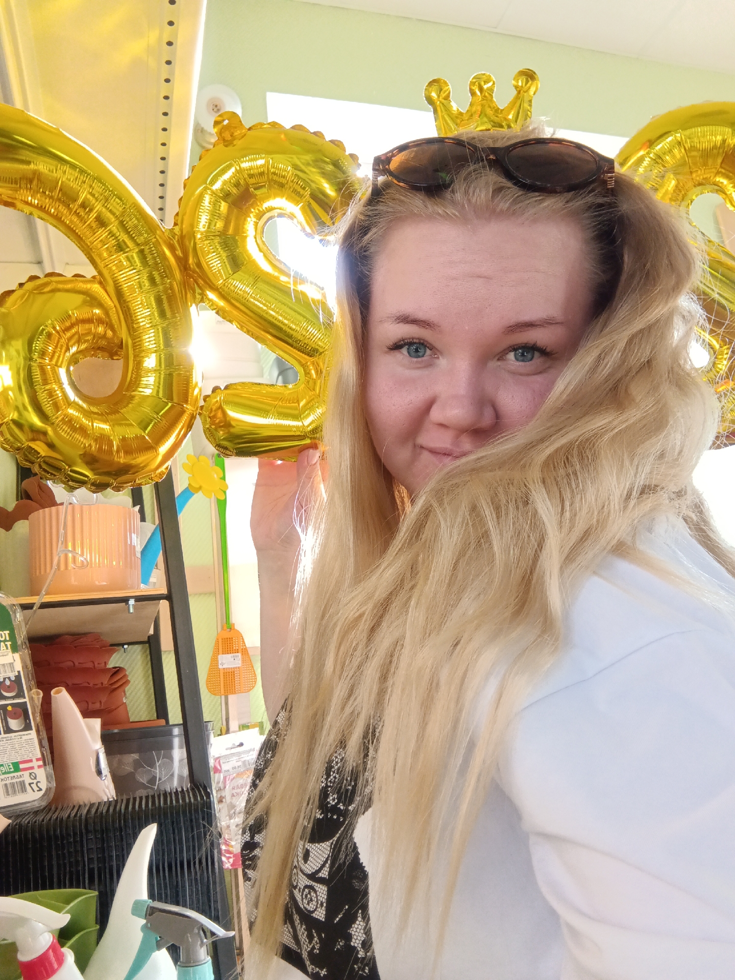 A selfie of a fair-skinned woman with blonde hair and blue eyes, wearing sunglasses on her head with a small gold crown behind them, holding two large gold balloons shaped like the numbers "62". The background includes a shelf with various items like a potted plant, cleaning supplies, and other decorative objects. The location is likely a shop or store, considering the merchandise displayed in the background. There is no verifiable information about the city of Фоки, Russia, present in this image.