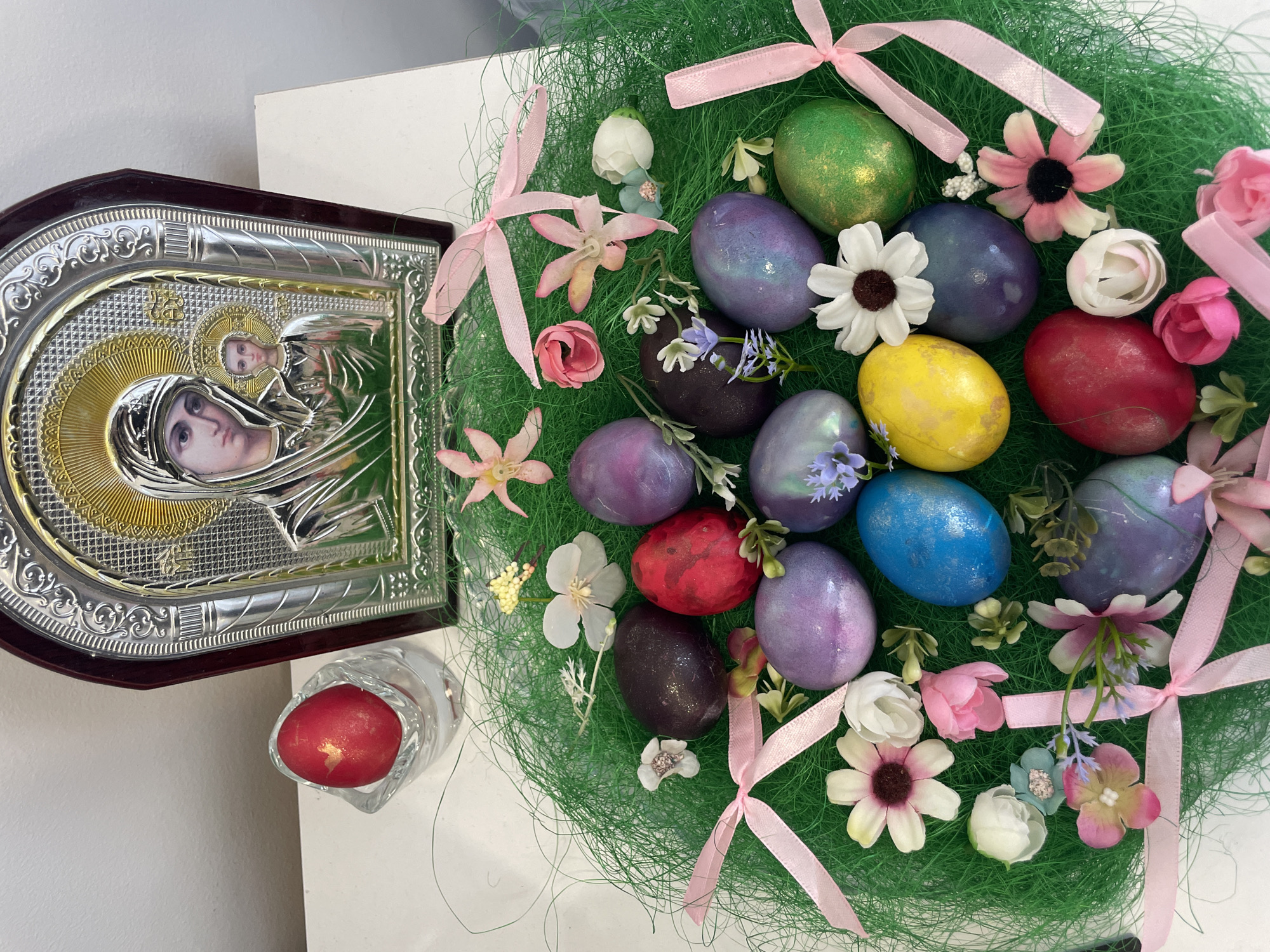 On a white surface rests an Orthodox icon, depicting a woman with a child, crafted with silver and gold plating and framed in dark wood. Beside it, a small glass holds a single red egg. Adjacent to these items is a circular Easter arrangement featuring green straw-like material, adorned with pastel-colored eggs (purple, blue, red, yellow, green) and artificial flowers in white, pink, and peach hues. The arrangement is accented with light pink ribbons tied into bows. The location is likely Varna, Bulgaria, given the prominence of Orthodox iconography and Easter egg traditions in that region.