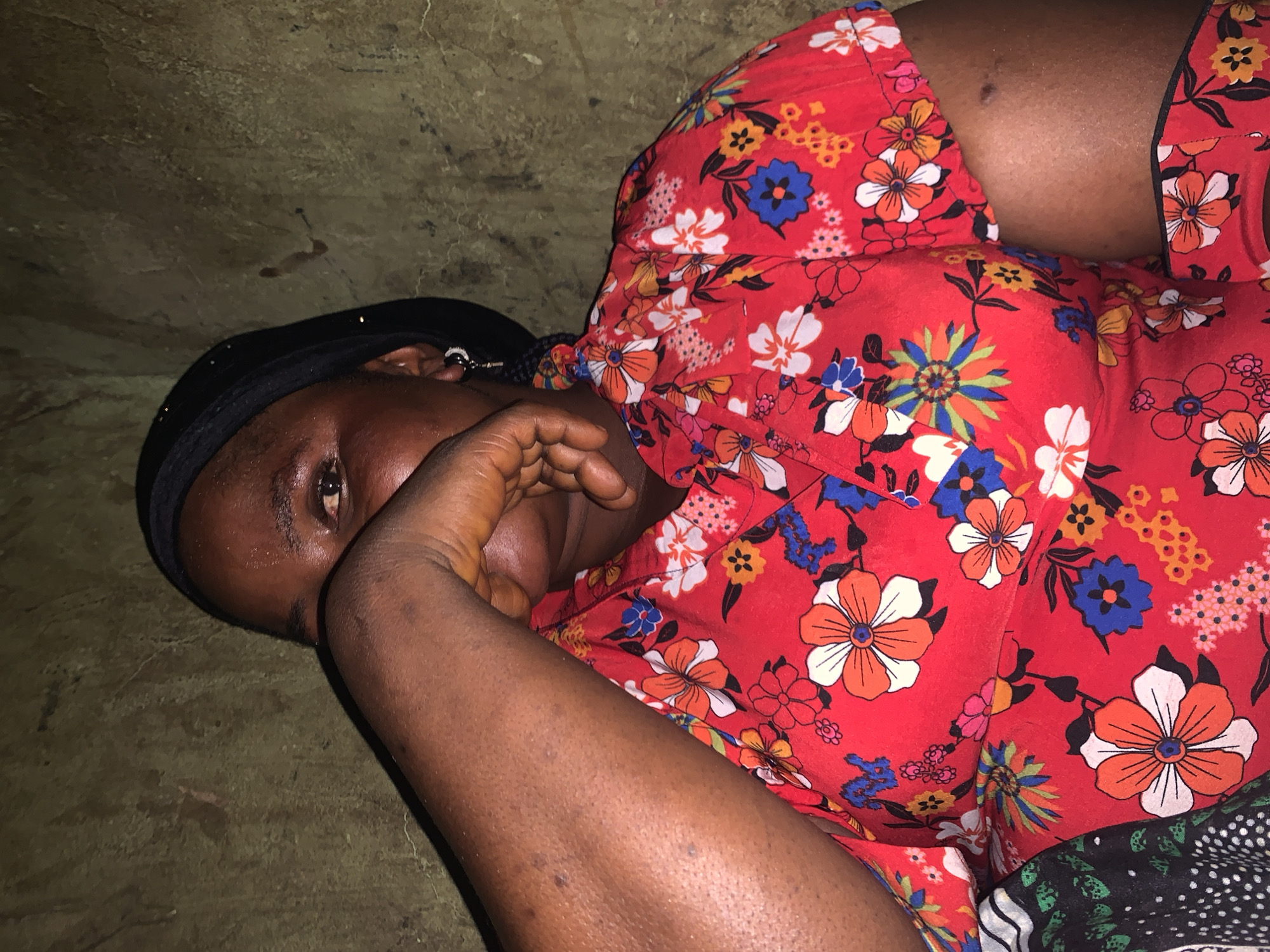 A person lies on a textured, earthy surface, their body angled towards the camera. They are wearing a vibrant red garment adorned with a colorful floral pattern of white, orange, blue, and yellow flowers. A black head covering is visible, framing their face. The person's right arm is raised, and their hand is positioned near their face, partially obscuring their features. One eye is visible, looking towards the camera, conveying a sense of weariness or introspection. The background is a rough, uneven surface, possibly dirt or stone, with some dark smudges. The lighting suggests an indoor setting or a shaded outdoor area, with no clear indication of the time of day or weather. The overall impression is one of quiet repose or perhaps discomfort. The context of "Project, Nigeria" provides a geographical and potentially thematic framework for the image. There is no visible text in the media file.