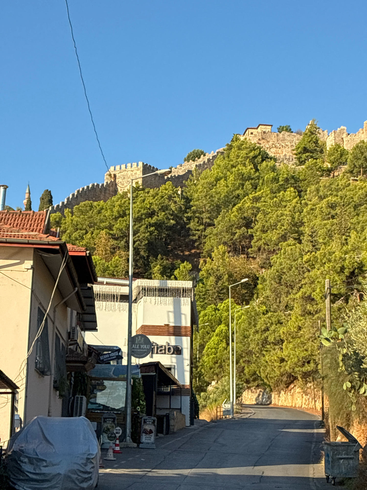 The image depicts a street scene in Alanya, Turkey, with a prominent castle wall on a hillside in the background. The castle wall, constructed of stone, stretches across the upper portion of the frame, topped with crenellations. It is surrounded by lush green trees, primarily pines, that grow densely on the steep slope. A clear blue sky forms the backdrop.
In the foreground, the street is paved with asphalt and appears to be a road leading uphill. On the left side of the street, a cream-colored building with a red-tiled roof is visible, featuring architectural details such as awnings and window frames. Next to it, a white building with multiple floors has signs for businesses, including one that reads "ALE YOLU RESTAURANT" and another with the word "fab." Further down the street, more shops and restaurants with outdoor seating are present, some with illuminated signs and posters.
Several objects are scattered along the street. A car is partially covered with a grey tarp on the left side. Streetlights are positioned along the road. There are also trash bins visible on the right side of the street, one of which is an open metal dumpster. A tall, slender minaret, characteristic of a mosque, is visible among the trees to the left of the castle wall. The overall impression is of a Mediterranean town with historical architecture and a vibrant street life. The lighting suggests it is daytime, with sunlight casting shadows on the road.
