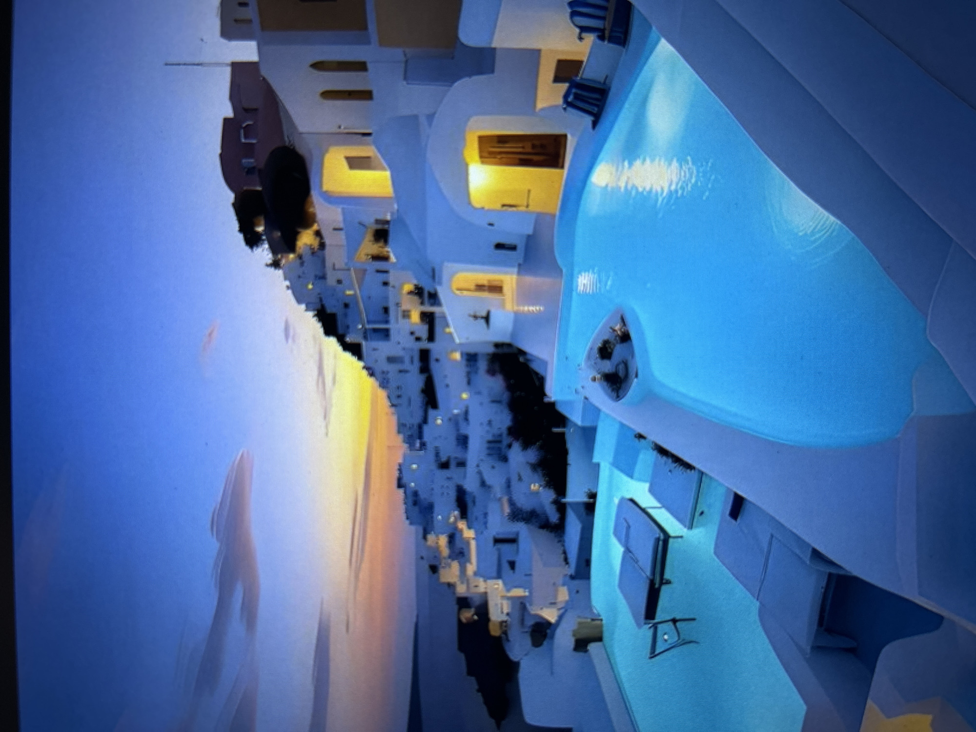 The image depicts a picturesque scene of a white, tiered village nestled on a hillside overlooking a body of water, likely the sea. The architecture is characterized by whitewashed buildings with curved roofs and arched doorways, many of which are illuminated with warm yellow light, suggesting it is either dusk or night. A prominent feature of the foreground is a large, irregularly shaped swimming pool filled with bright blue, illuminated water. Several lounge chairs and tables are visible around and within the pool area, some submerged. In the distance, the sky displays a gradient of colors, transitioning from deep blue at the top to soft orange and yellow hues near the horizon, indicating a sunset or sunrise. Clouds are present, some appearing wispy and elongated. The overall impression is one of a luxurious resort or hotel complex, possibly in a Mediterranean setting, given the architectural style.
The image does not contain any elements that would identify it as being located in the city of Burgas, Bulgaria. The architecture and landscape are more consistent with popular tourist destinations like Santorini, Greece.