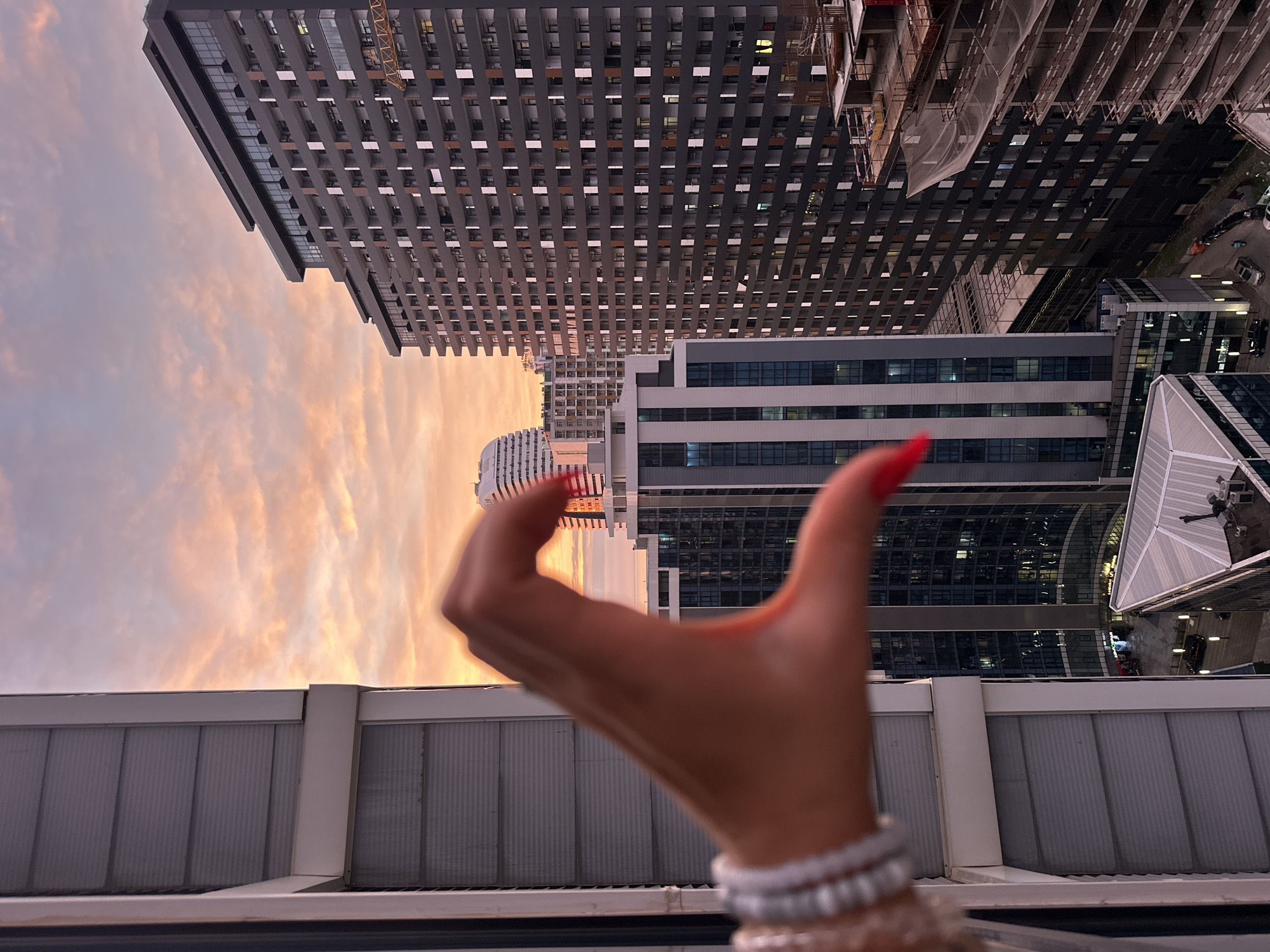 The image captures a cityscape at dusk with a focus on a hand in the foreground making a heart shape. The hand, with bright red painted nails and a beaded bracelet, is positioned to the left of the frame. The background is dominated by tall, modern buildings, some appearing to be under construction. The sky displays a warm, gradient of orange and pink hues, indicative of sunset.
The setting appears to be urban, likely from a high vantage point such as a balcony or rooftop, overlooking a city. The buildings suggest a developed area with a mix of residential and commercial structures. The lighting conditions imply it is either late afternoon or early evening. No specific text is visible in the image. The overall mood seems relaxed and appreciative of the view.