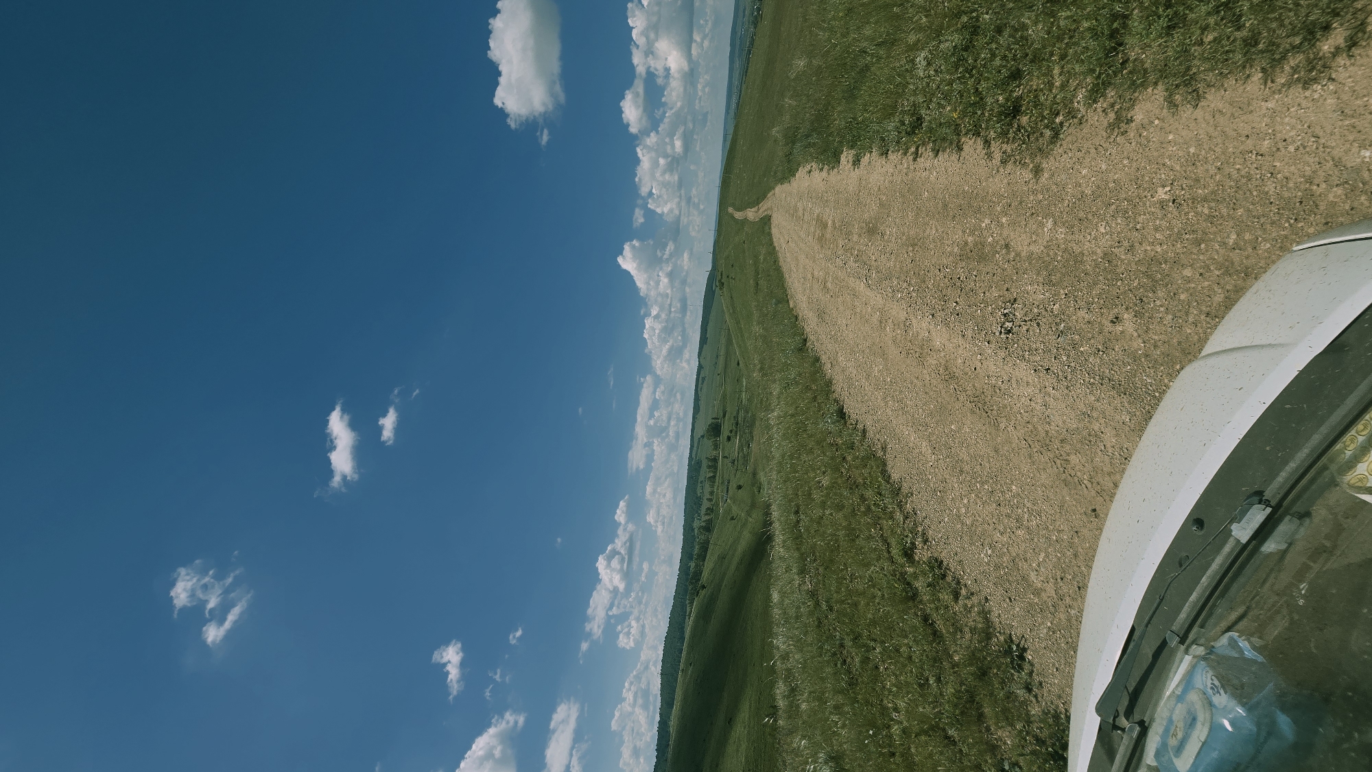 The image depicts a dirt road winding through rolling green hills under a bright blue sky with scattered white clouds. The perspective is from inside a white vehicle, showing the hood and windshield in the foreground. The landscape appears rural and undeveloped, with grassy slopes and distant trees. The location is identified as Kudashevo, Russia. There are no people or specific actions visible, only the environment and the car.