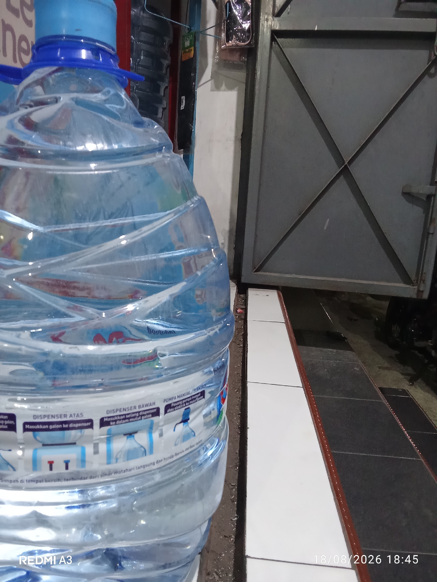 A close-up, eye-level, outdoor shot captures a large plastic water bottle prominently in the foreground, with a metal garage door and a tiled floor in the background. The water bottle, which appears to be a 19-liter refillable type, is transparent and filled with clear water, showing subtle indentations and reflections on its surface. A blue plastic cap with a white inner seal is visible at the top.
The background features a grey, industrial-style metal garage door with a diamond pattern formed by diagonal beams. The door is partially ajar, revealing a dark interior. To the left of the door, a white wall is visible. The foreground ground is covered in a tiled pattern, with alternating sections of white and dark grey tiles, bordered by a thin red strip.
Text labels are present on the water bottle, though mostly illegible due to image resolution and focus. One label appears to contain instructions for use with a dispenser, depicting illustrations of a top dispenser and a bottom dispenser, as well as a manual pump. The bottom left corner of the image displays "REDMI A3" indicating the phone model used. The bottom right corner shows a timestamp: "18/08/2026 18:45". The setting appears to be an urban environment, possibly a shop or a service area, given the presence of the garage door and water dispenser instructions. The lighting is dim, suggesting it is either evening or a poorly lit area.