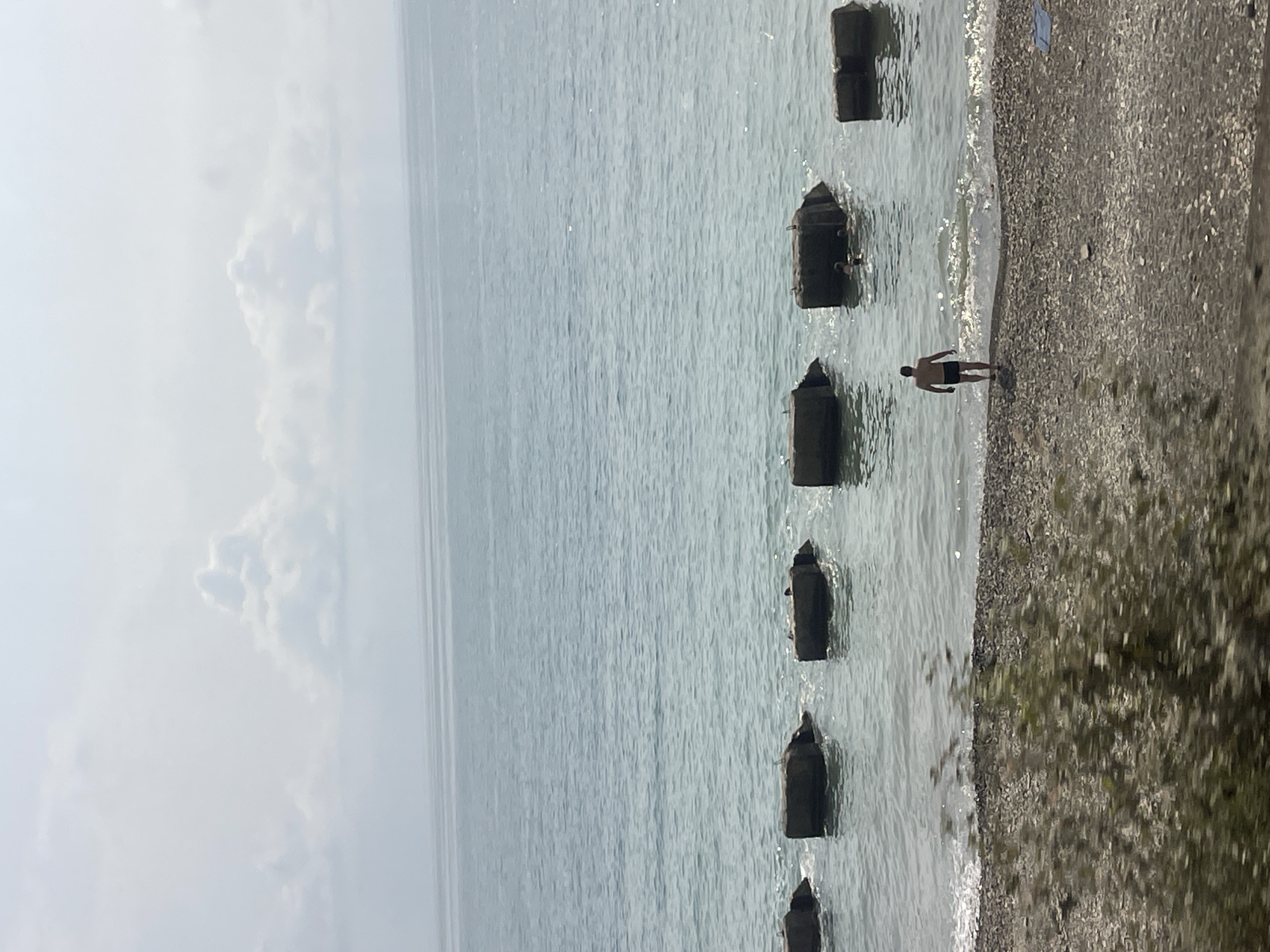 In Sochi, Russia, on a pebbled beach, a lone man stands facing the calm, blue sea. The man, wearing black shorts, is positioned at the water's edge where gentle waves break. Further out in the water, several large, submerged concrete blocks act as breakwaters or submerged structures. Some of these blocks have birds perched on them. In the distance, more concrete blocks are visible in the water, and a small group of people are swimming near one of them. The sky is overcast with soft, scattered clouds. The overall scene depicts a peaceful coastal environment.