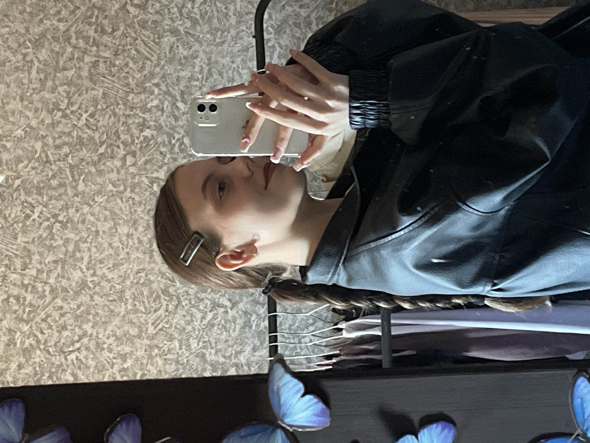 A woman is taking a selfie with her phone in front of a textured wall. She is wearing a black leather jacket. Her hair is braided and has a silver hair clip. In the foreground, there are decorations of blue butterflies on a black surface. The location details are not present.