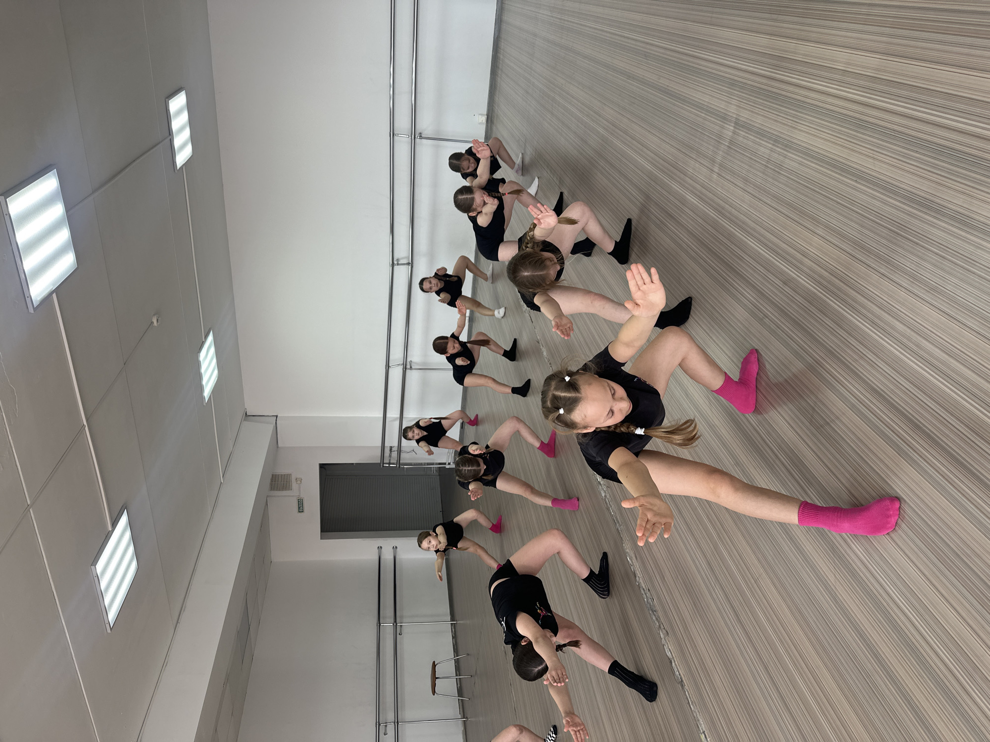 A group of approximately ten young girls are in a dance studio in Votkinsk, Russia. They are all wearing black dance attire, and some have on colored socks. The girls are arranged in a line, executing a synchronized dance move with their legs spread and arms extended. The dance studio has a ballet barre on the left side, and the floor is light-colored with a linear pattern. The room has a white ceiling with rectangular fluorescent lights. The studio appears well-lit and clean. An exit door is visible in the background, adjacent to the ballet barre.