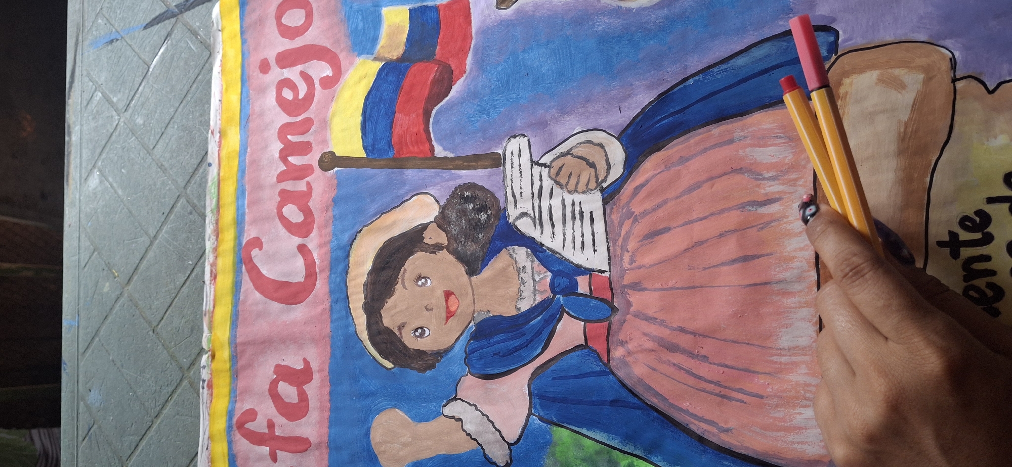 A handmade poster, likely painted, features a woman in historical clothing, possibly a depiction of a figure from Venezuelan or Colombian history. She holds what appears to be musical notes or a small book. Behind her is the flag of Colombia. The backdrop is painted in pink, blue, and purple. The name "fa Canejo" is written in large red letters along the left side of the poster. A portion of a word ending in "ente de" is visible on the right. A human hand holds two pens, one orange and one pink. The background appears to be a textured grey surface. The exact location in Vallecito, Venezuela is not specified by visual information.