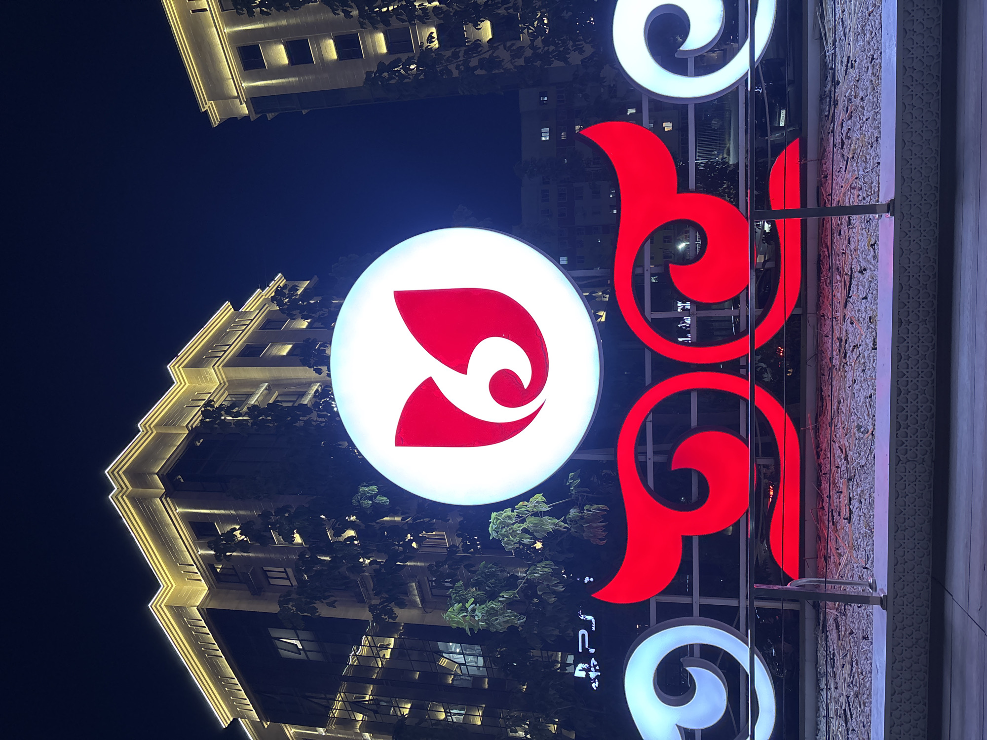 The image captures a nighttime view of a plaza in Shymkent, Kazakhstan, likely in a commercial or entertainment district. Prominently featured is a large, illuminated circular sign with a red, stylized emblem resembling a blooming flower or a folded petal. Below this, a vibrant, large-scale decorative element is displayed, composed of two symmetrical, swirling shapes – one in bright red and the other in white. These illuminated structures are set against a backdrop of modern, multi-story buildings that are lit from within and have decorative external lighting along their facades, highlighting their architectural details. Trees with visible foliage are interspersed between the buildings and the signage. In the lower left corner of the image, a smaller illuminated sign with the letters "PSJ" is visible. The foreground shows a decorative border with a patterned design and a surface covered in white stones or gravel, with a metal railing running across it. The overall scene suggests a lively urban environment, possibly a public space designed for evening gatherings or advertising.