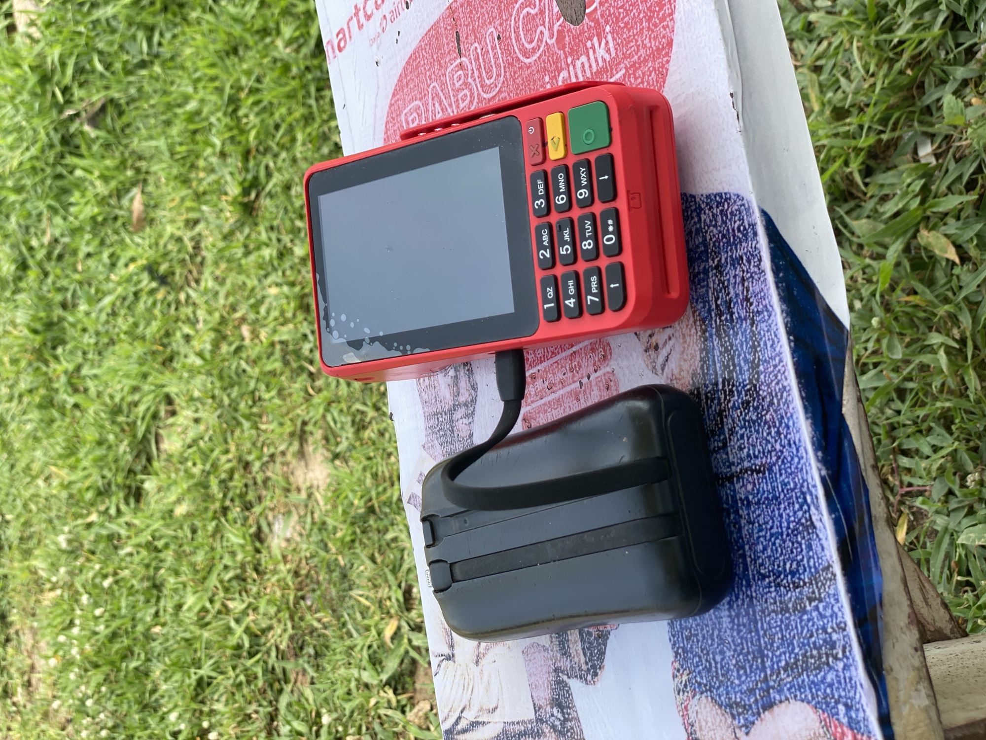 An overhead shot shows a red point-of-sale device with a black accessory on its left, both resting on a bench. The bench is positioned on a patch of green grass, with more grass and some dirt visible in the background. The point-of-sale device has a screen and a keypad with numbers and letters. The black accessory is rectangular and appears to be a power bank or similar device, with a strap attached to it. The bench is decorated with a printed image of a woman's face. The scene is outdoors, suggesting it may be in a park or public area in Jalingo, Nigeria.