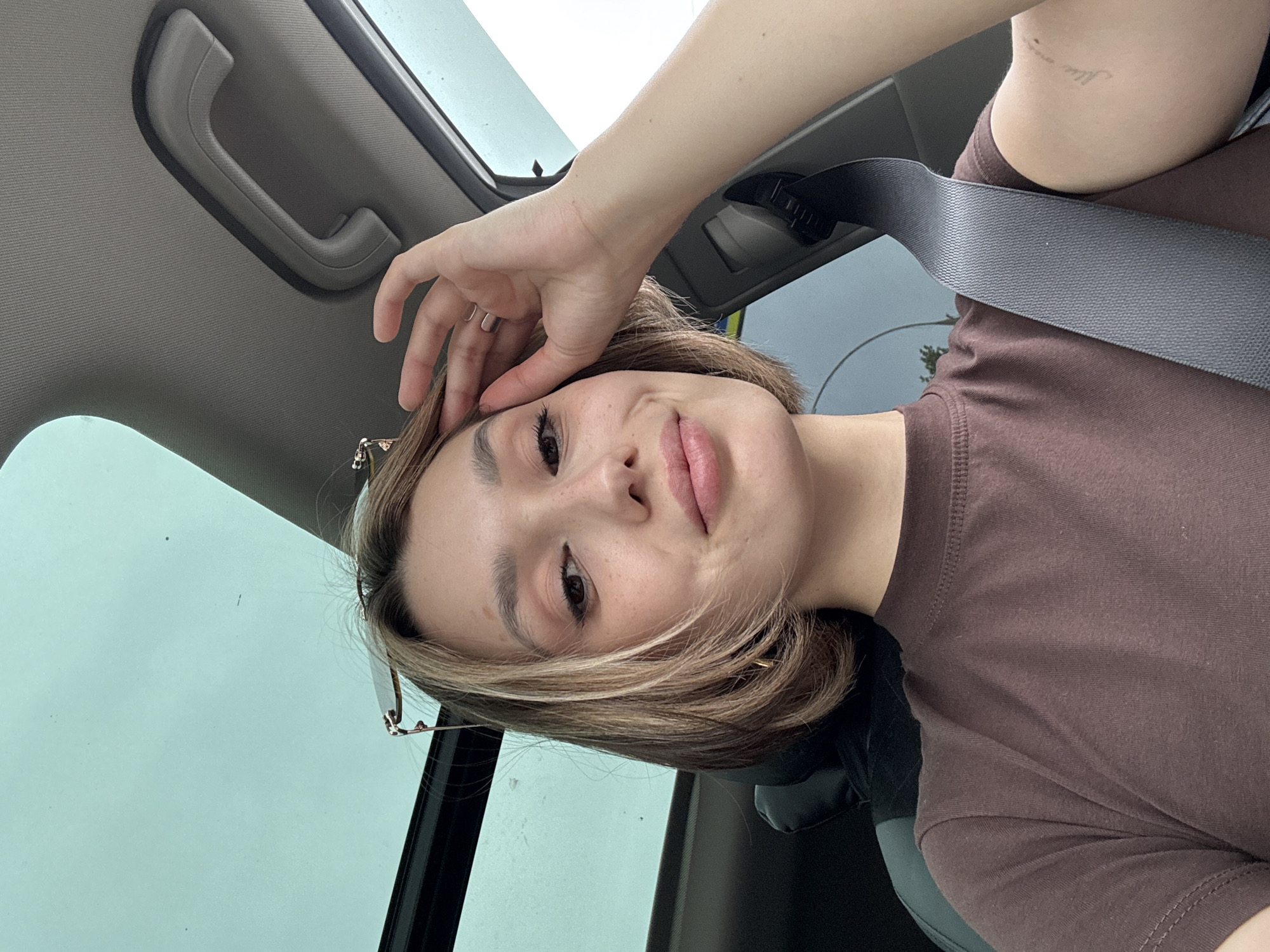 A young woman with short blonde hair and light makeup is in the driver's seat of a car, looking at the camera. She is wearing a brown t-shirt and has her left hand on her forehead, touching her hair. A seatbelt is visible across her chest. The car's sunroof is open, and the sky is visible above. The background is blurred, but hints of trees and a building suggest an urban or suburban setting.