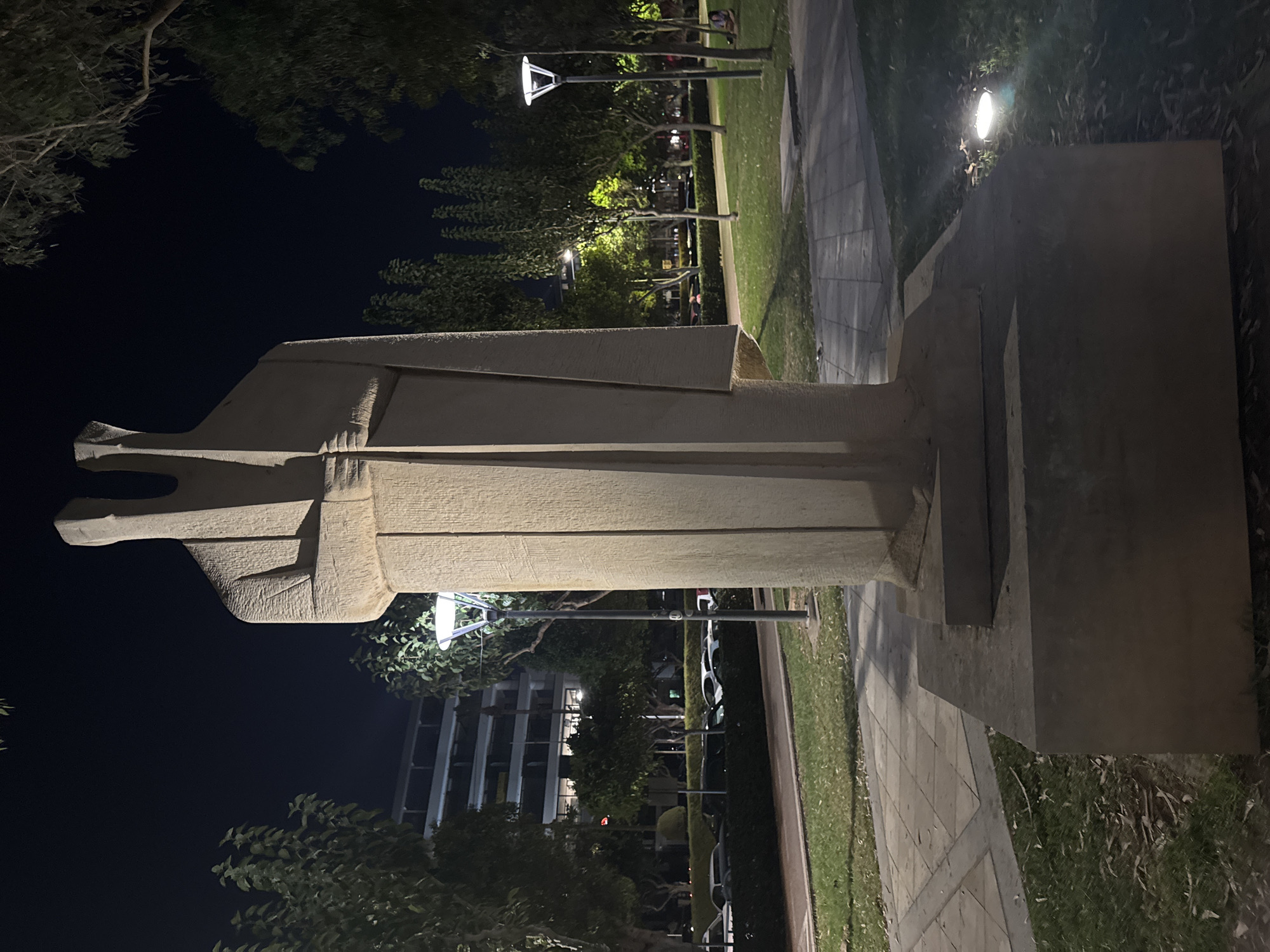 The image shows a large, abstract stone sculpture of two figures standing side-by-side, illuminated by lights at night. The sculpture is located in a park or public space in Limassol, Cyprus, likely near the city center or a coastal area, given the presence of trees and a well-maintained landscape. In the background, there is a multi-story building with illuminated windows, suggesting an urban environment. Cars are parked along a road in the middle ground, and streetlights are visible, casting a glow on the surroundings. The ground is paved with tiles and covered with grass and fallen leaves. The overall atmosphere is serene and quiet, typical of a public space at night.