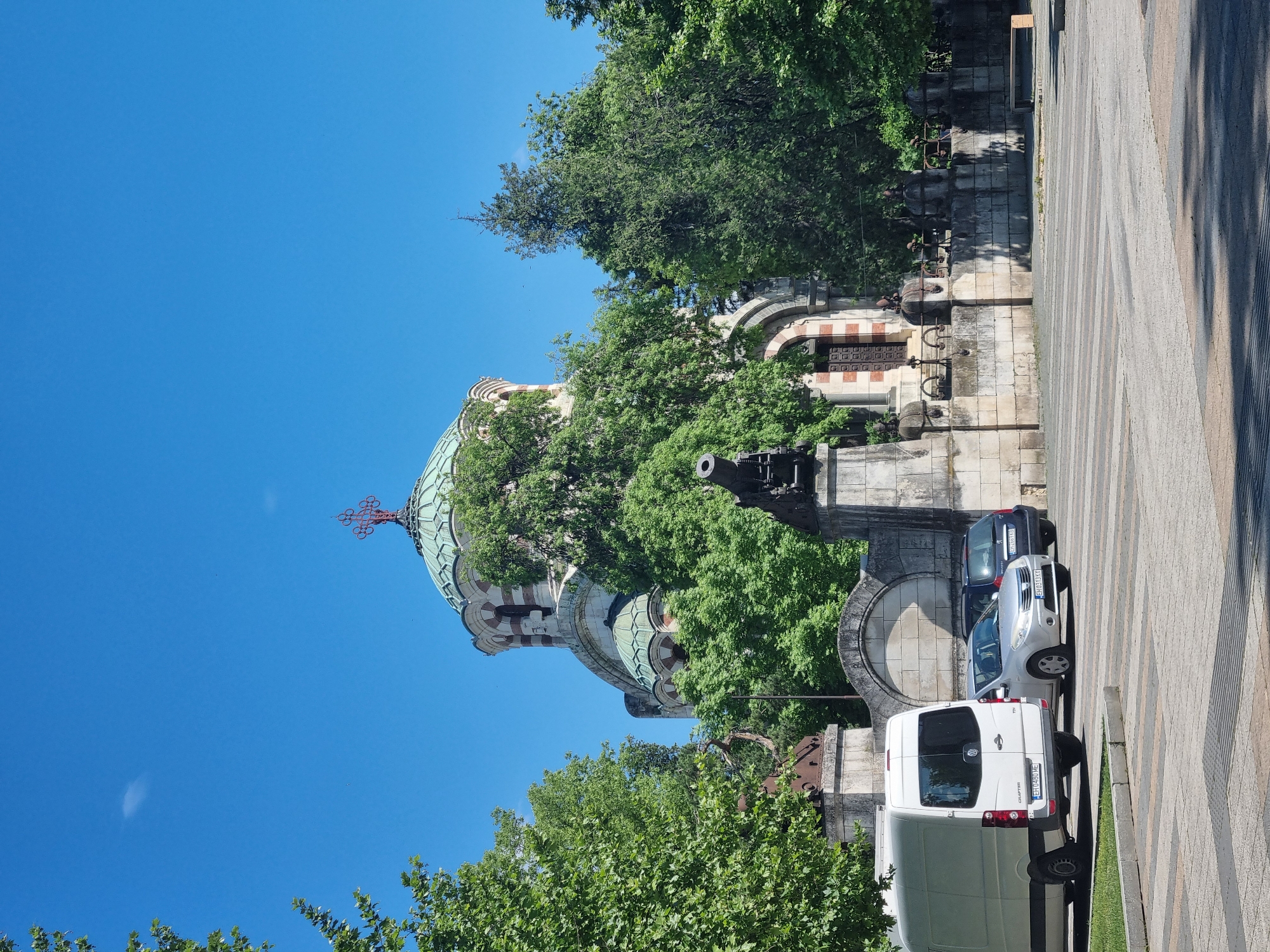 Here is a description of the image:
The image shows the St George the Victorious Chapel Mausoleum in Pleven, Bulgaria. The dome of the chapel rises into a clear blue sky. It's partially obscured by trees. A stone wall with arches supports a cannon. Parked on the cobblestone street are a white van, a silver car, and a dark-colored car. The photograph is taken from a low angle.
