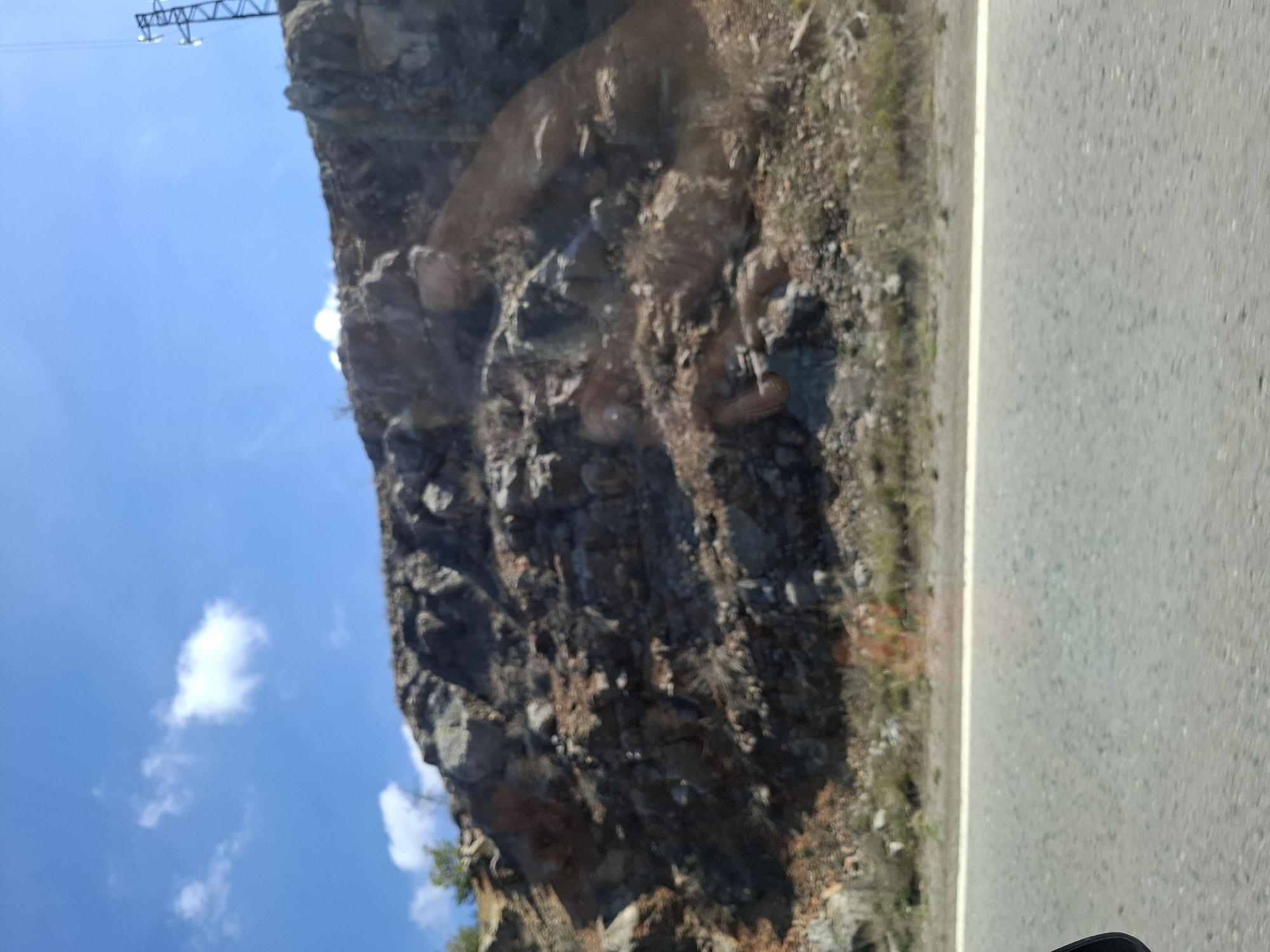 The image depicts a view from inside a vehicle, likely moving along a paved road. To the left of the road is a rocky hillside or cliff face covered with vegetation. The sky is visible above the hillside, with some clouds present. An electrical transmission tower is seen at the top of the frame. The location is possibly near Ponor, Bulgaria.