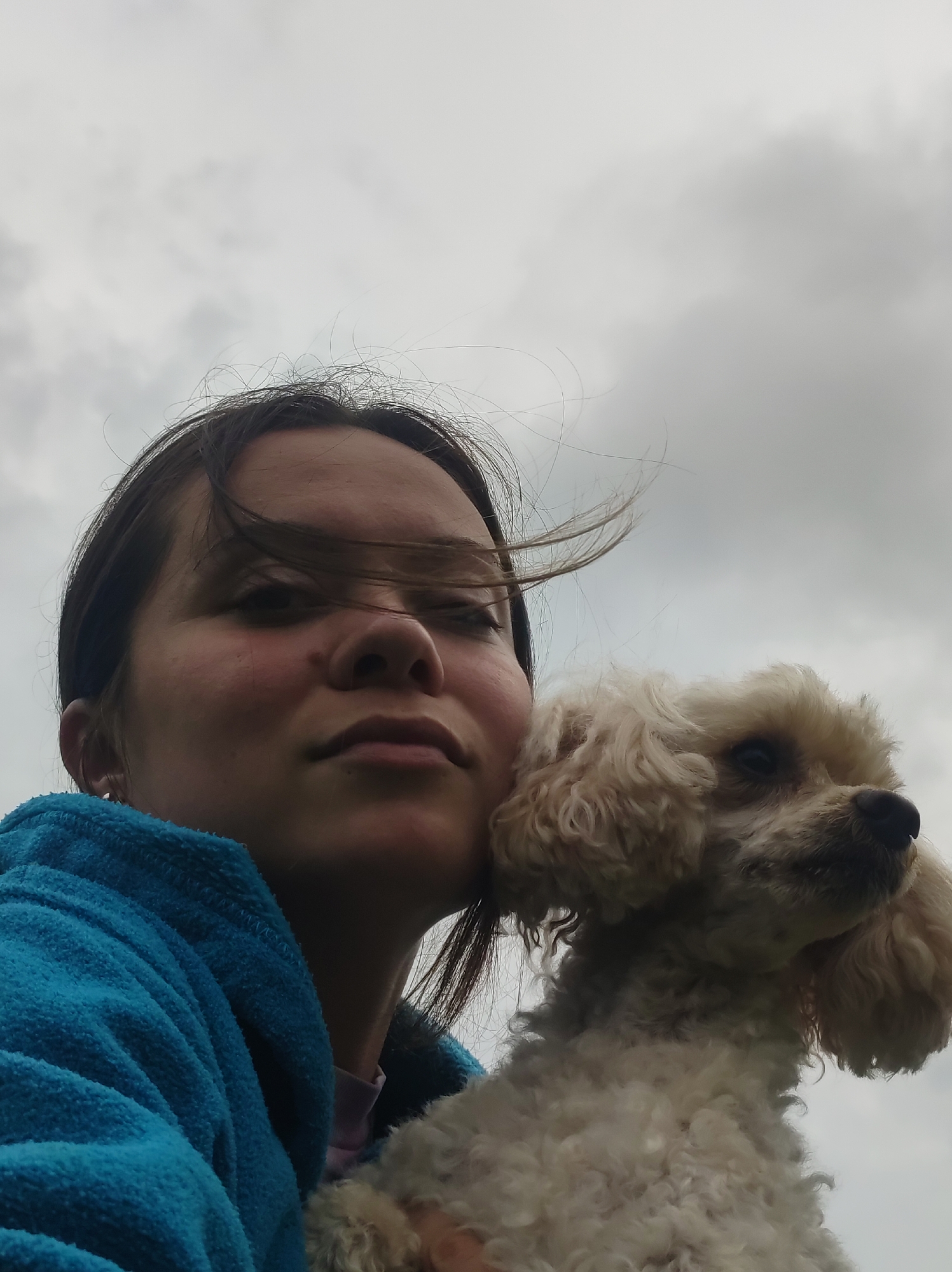 A young person is holding a light-colored poodle against a cloudy sky. The person is wearing a blue hooded garment and has dark hair blowing in the wind. The poodle's head is tilted upwards. The image is a close-up shot, with the person's face and the poodle occupying most of the frame. The background is an overcast sky. The scene is outdoors, and no specific urban features are visible to identify it as being in Фоки, Russia.