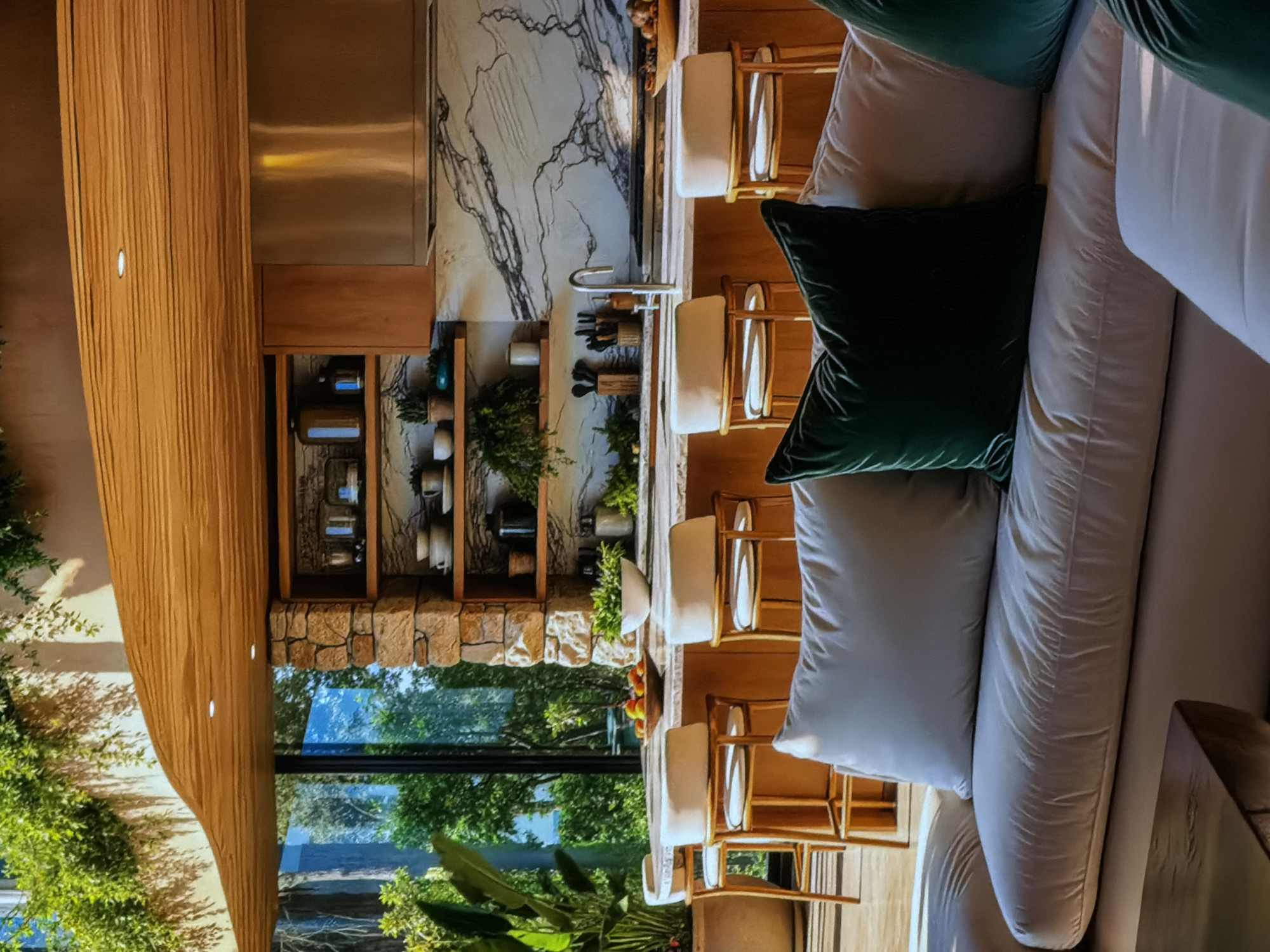 This image captures the interior of a luxurious and modern living space, likely located in Sofiivska Borshchahivka, Ukraine. The scene is illuminated by natural daylight streaming through large windows, suggesting a clear day.
In the foreground, a comfortable, light-colored sectional sofa occupies the right side of the frame. It is adorned with several cushions, including a prominent dark green velvet throw pillow and lighter grey/beige pillows.
Beyond the sofa, a sleek kitchen island or bar area extends across the midground, featuring at least four modern bar stools with light-colored upholstered seats and light wooden frames. This area serves as a focal point, separating the lounging space from the kitchen.
The kitchen, situated in the background, boasts striking white marble countertops and backsplash with distinctive dark veining. A modern stainless steel appliance, possibly a range hood, is visible above a counter. Below, a contemporary kitchen sink with a high-arc faucet is surrounded by various kitchen utensils, suggesting functionality. Open wooden shelving units are integrated into a stone wall section, displaying neatly arranged dinnerware, jars, and small potted plants, adding a touch of domesticity and greenery to the space.
A long, curved wooden feature, possibly a suspended ceiling element or a decorative beam with integrated lighting, spans the left side of the image, contributing to the organic and warm aesthetic. Through the expansive windows, lush green foliage and trees are visible, blurring the lines between indoor and outdoor living and enhancing the serene atmosphere. No people are present, and no specific activity is occurring, allowing the focus to remain on the architectural design and tranquil ambiance. The overall impression is one of sophisticated comfort and a strong connection to nature.