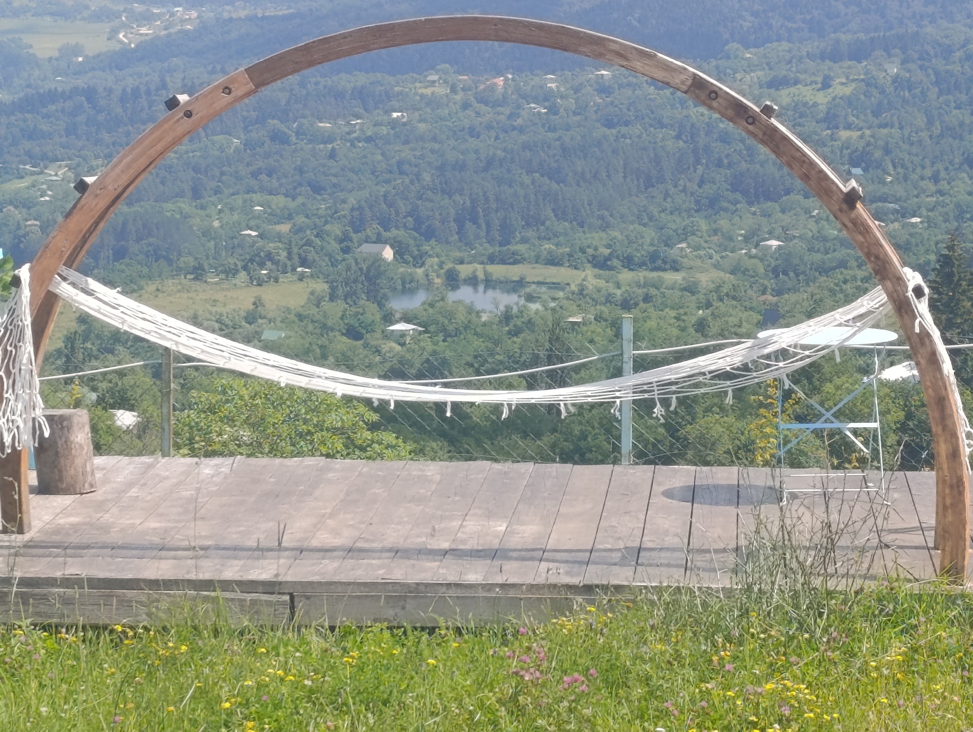 The image depicts a wooden archway with a white net hammock strung beneath it, positioned on a wooden deck overlooking a verdant valley. The valley is densely forested with scattered houses and a small body of water, possibly a lake or pond. In the foreground, there is a patch of green grass dotted with small yellow and pink wildflowers. A tree stump is visible to the left of the hammock, and to the right, a metal table and chair are partially obscured by the hammock and surrounding foliage. The overall scene suggests an outdoor recreational area, likely in a rural or semi-rural setting in or around Nikortsminda, Georgia, given the characteristic landscape and architecture of the region. The lighting indicates it is daytime with bright sunlight.