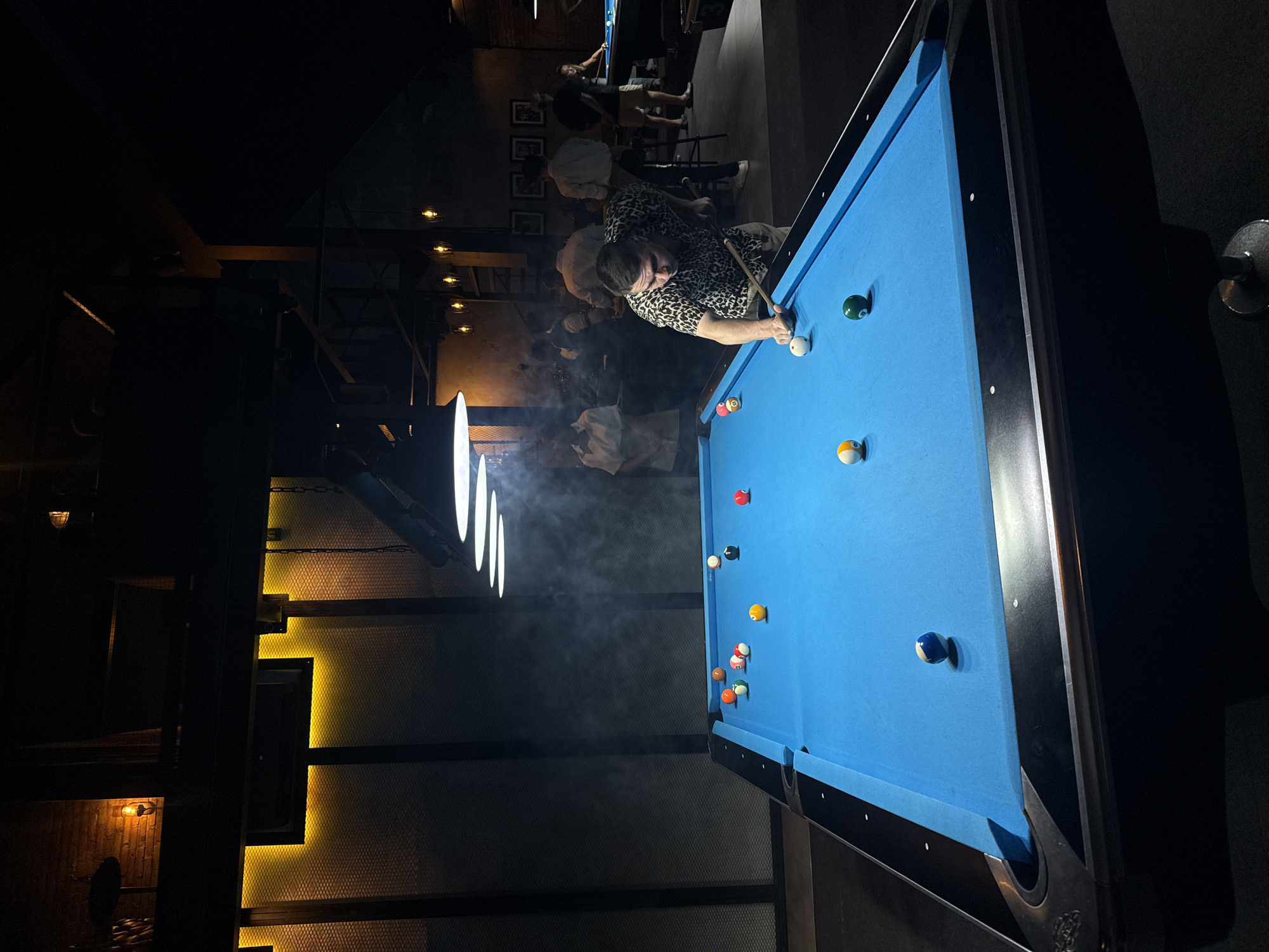 This media file captures an indoor, dimly lit scene, likely a pool hall or bar, with a strong focus on a game of billiards.
The main subject is a man, positioned on the right side of the frame (when correctly oriented), actively playing pool. He wears a distinctive short-sleeved leopard-print shirt and light-colored pants, leaning intently over a vibrant blue pool table. He is holding a cue stick, poised to strike the white cue ball. His expression suggests concentration on the game.
The pool table itself is covered in bright blue felt, contrasting sharply with the dark surroundings. Scattered across its surface are multiple colorful billiard balls, including solids, stripes, and the cue ball, indicating a game in progress. The table features dark, possibly black, rails and pockets.
In the background, several other people are visible, though mostly out of focus due to the low lighting and shallow depth of field. They appear to be casually standing or sitting, observing the game or engaged in their own conversations. Another pool table with a player can be discerned further back in the room.
Notable details include the atmospheric lighting. Bright white strip lights hang directly above the main pool table, casting a strong illumination on the playing surface and interacting with visible wisps of smoke or haze in the air. Complementing these are warm, yellowish-orange accent lights that highlight various architectural elements, such as wall panels, structural beams, and perhaps decorative fixtures, giving the venue an industrial yet cozy ambiance. The walls exhibit a textured pattern, resembling mesh or a similar material. The setting suggests an evening or nighttime activity. No visible text can be clearly identified.