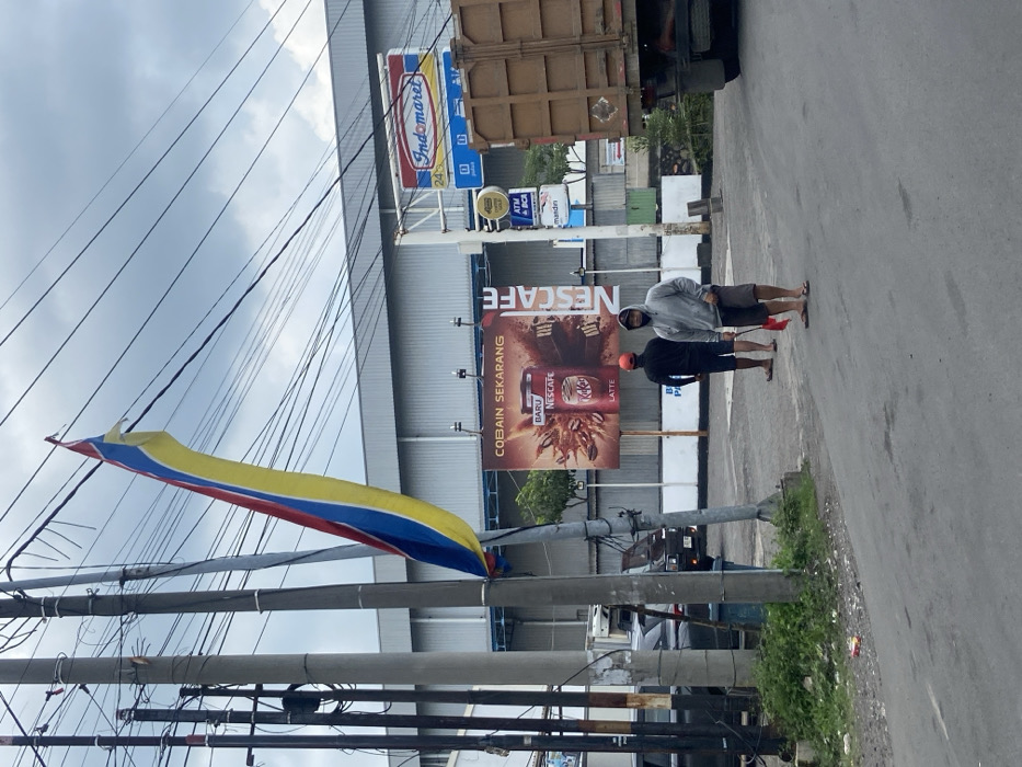A sideways street scene shows an Indomaret store with a Nescafe advertisement. Two men stand on the sidewalk by the sign, one in a hooded sweatshirt and shorts, the other in darker clothing. A truck is partially visible behind them. In the foreground, power lines crisscross, and a yellow, red, and blue flag hangs from a pole. The sky is overcast.