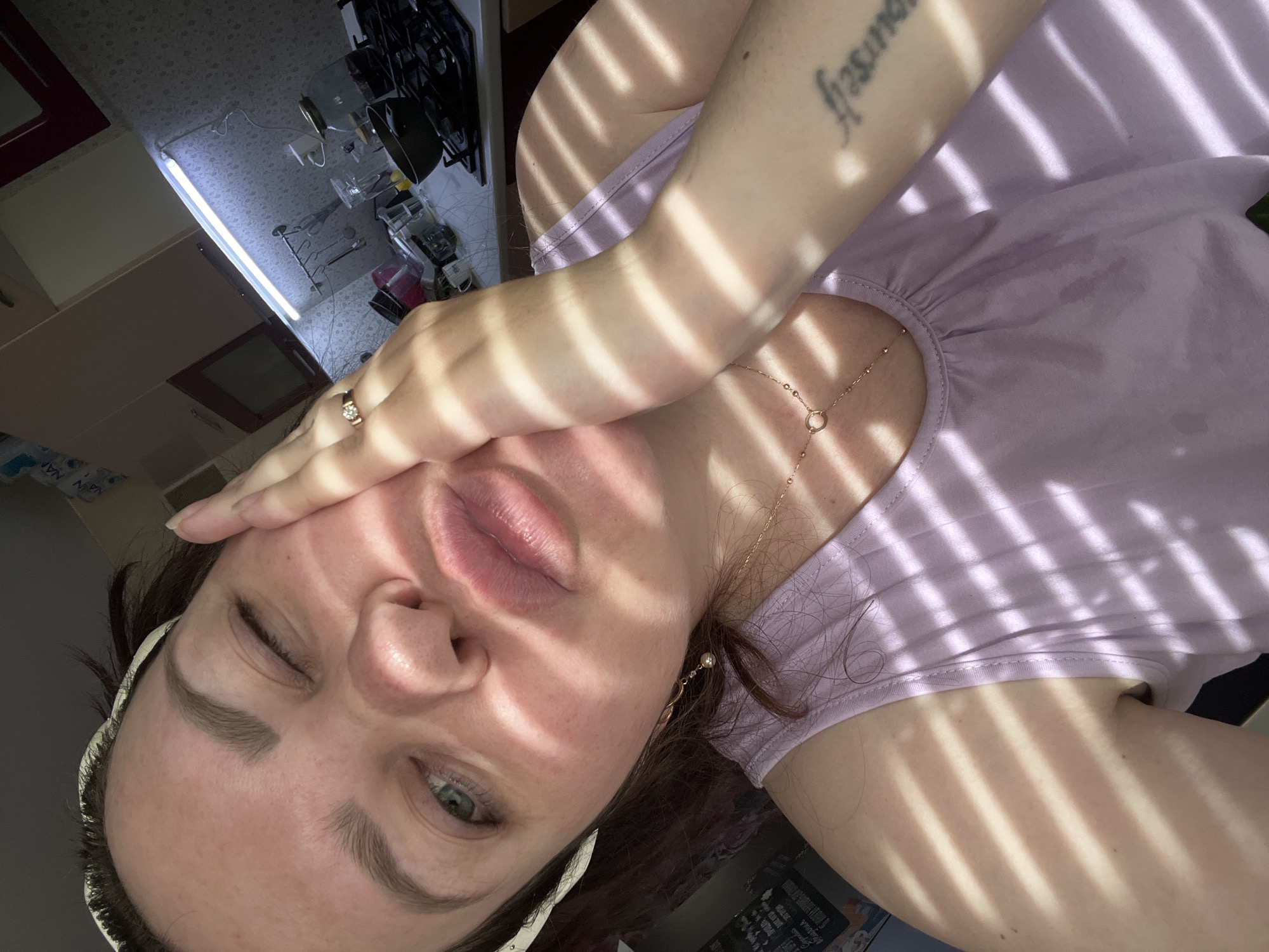 A close-up, angled selfie photograph shows a Caucasian woman indoors with light and shadow stripes across her face and upper body. She is wearing a light purple tank top and a white headband. A gold necklace and earring are visible. She has a ring on her finger. Her left eye is open, and her right eye is closed in a wink. Her hand is touching her forehead. A tattoo is visible on her right arm. The background includes a kitchen area with cabinets, a stovetop, and fluorescent lighting. There is no evidence to support that the image was taken in Noviy, Russia.