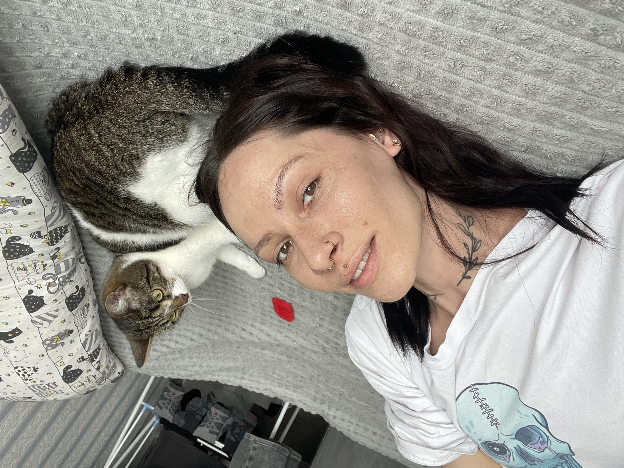 A medium, eye-level, close-up shot depicts a woman and a cat lying on a grey textured surface resembling a bedspread. The woman is positioned on the right side of the frame, with dark brown hair, fair skin, and a white t-shirt with a blue and white skull graphic. She has facial piercings on her nose and ear, and a small, dark tattoo on her neck. She is looking towards the camera, with a slight smile.
The cat is lying to the left of the woman. It has a brown tabby head with green eyes and white chest and belly, and a striped brown tail. A pillow patterned with cartoon cats is beside the cat.
The background includes a glimpse of what appears to be clothing on a rack. A small red object, possibly a toy, sits between the woman and the cat. The location is not identifiable as Izhevsk, Russia; it appears to be an interior setting.