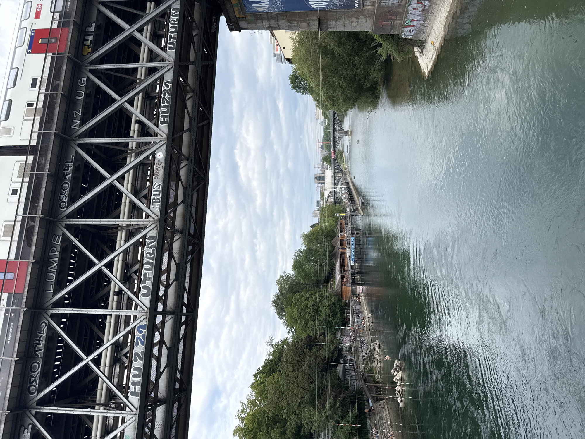 The image shows a view from a bridge overlooking a river in Zurich, Switzerland. On the left, a train with the markings "SBB CFF FFS" is visible, partially obscured by the bridge's metal structure which is adorned with graffiti. Text visible on the train includes "04 1852 8 602 208-3". Graffiti on the bridge structure includes words like "LUMPE", "OSLO THL", "NZUG", "HUZZA", and "UTURN".
The river is a wide expanse of green water flowing beneath the bridge. On the right bank of the river, there are trees and buildings. Further along the river, another bridge can be seen in the distance. The riverbank is lined with people, some of whom appear to be swimming or sunbathing. The scene suggests a warm, possibly summer, day with scattered clouds in the sky. The atmosphere seems relaxed and recreational, with people enjoying the river on a sunny day. The visible text and the SBB train suggest a Swiss setting.