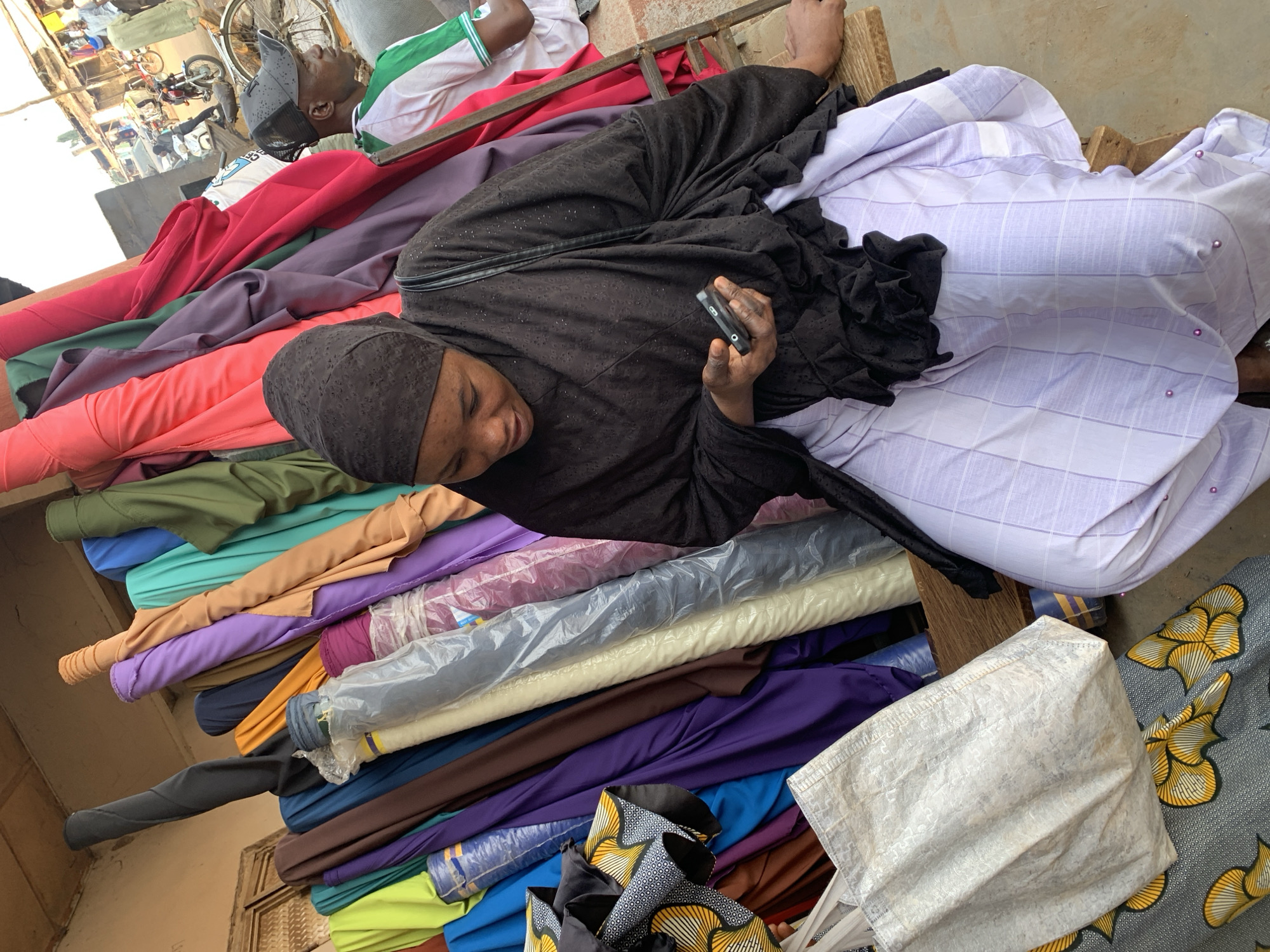 The image depicts a woman in Kafanchan, Nigeria, amidst a vibrant textile market. She is lying on a stack of colorful fabric rolls, using a mobile phone. The textiles vary in color and pattern, ranging from solid hues to patterned designs. Other people are visible in the background.