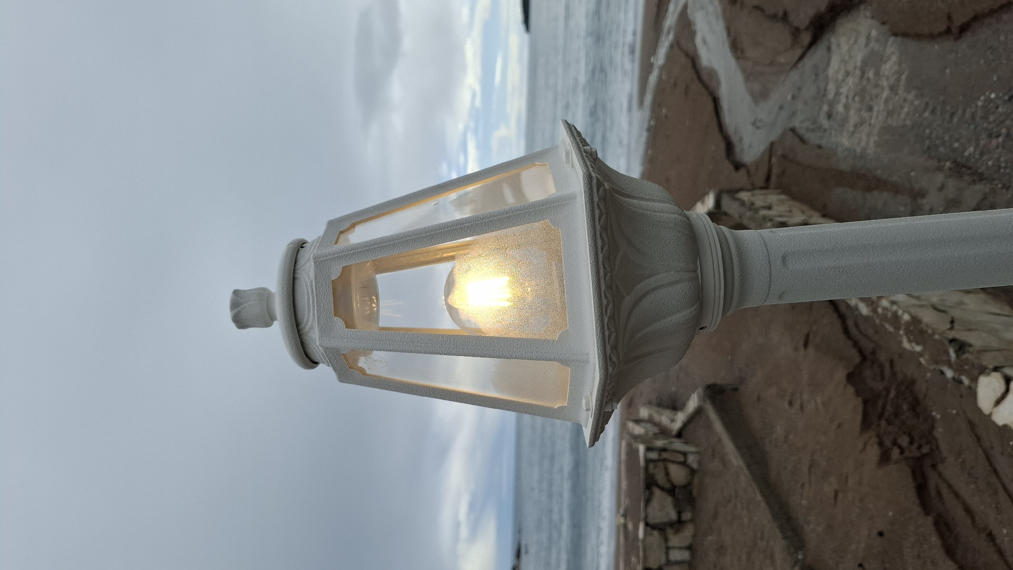 The image displays an illuminated white ornamental lamp situated on a coastal area in Budva, Montenegro. The lamp occupies the foreground, featuring a hexagonal lantern head with clear panels encasing an active LED filament bulb, which emits a warm, bright light. The lamp's exterior possesses a granular texture, and its uppermost finial resembles a closed flower bud. The base of the lantern head is adorned with sculpted, leaf-like patterns, and the visible section of the lamp pole is cylindrical with vertical fluting.
The immediate ground behind the lamp consists of reddish-brown sediment, likely sand or earth, marked by a distinct, narrow water channel winding towards the sea. To the lower left, a partial view of a low wall constructed from irregularly shaped stones is visible. The midground is dominated by the sea, which appears calm with minimal surface ripples. The horizon line separates the sea from a predominantly overcast sky, characterized by varying shades of grey and white cloud formations. A dark landmass is discernible on the distant right horizon, with a less defined landmass present on the far left. No individuals are depicted in the scene. The operational lamp indicates conditions of reduced ambient light.