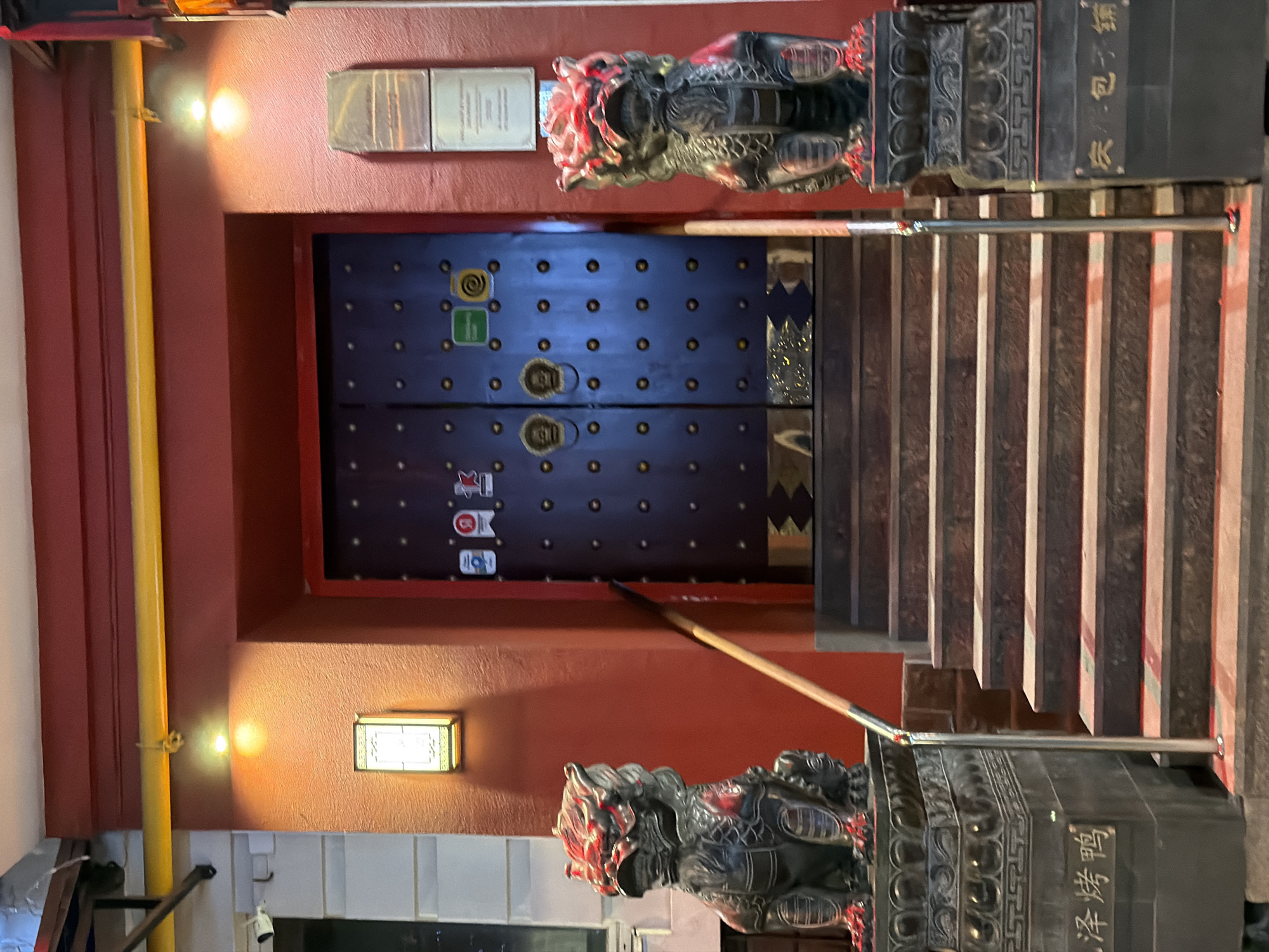 The image depicts the entrance to a building in Moscow, Russia, likely a restaurant given the signage and decorative elements. Dominating the scene are a set of dark wooden double doors, adorned with numerous small golden studs and two ornate brass door knockers. Flanking the entrance are two detailed stone lion statues, known as "foo dogs" or "guardian lions," each perched atop a decorative pedestal. The lion on the left has its mouth open, while the one on the right has its mouth closed, a common representation. The pedestals are inscribed with Chinese characters.
The entrance is accessed by a wide, stone staircase with multiple steps. A metal handrail runs along the right side of the stairs. The building's facade is painted a deep red, with decorative yellow piping running above the entrance. Two wall-mounted light fixtures illuminate the area, casting warm light onto the textured red wall. On the left side of the entrance, a security camera is visible mounted on a white building facade. Above the red wall, a glimpse of a bright yellow pipe is seen. The overall impression is one of a building with a strong Asian cultural influence, likely a Chinese restaurant, situated in an urban environment. There are no people visible in the image.