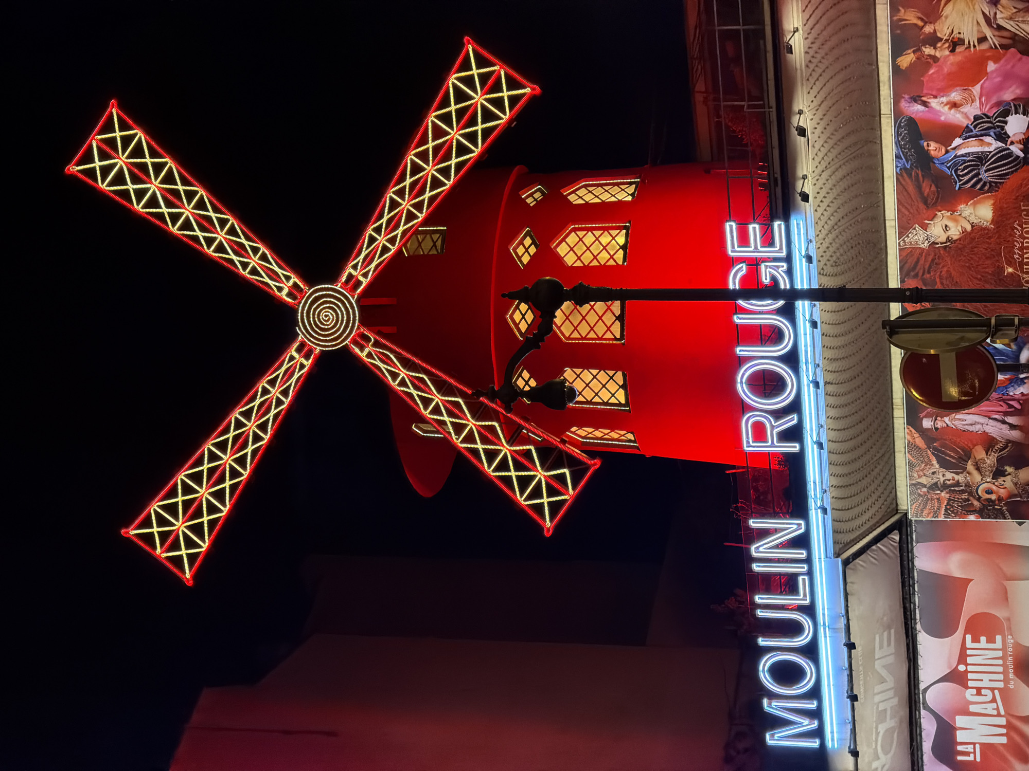 The image captures the Moulin Rouge in Paris, France, at night. The iconic red windmill is illuminated with glowing yellow lights outlining its sails and windows. A blue neon sign spells out "MOULIN ROUGE" vertically. To the right of the sign, posters with images of performers are displayed. The surrounding environment is dark.