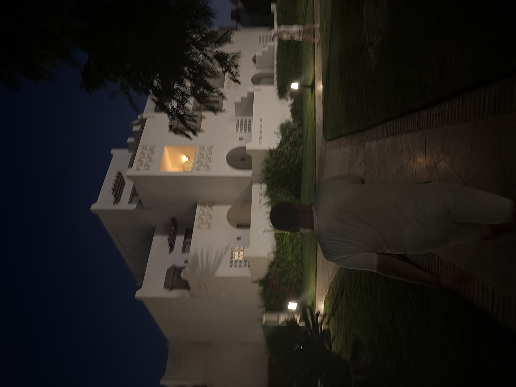 The image depicts a nighttime scene in Hurghada, Egypt, likely within a resort complex given the architecture. In the foreground, a person wearing a white t-shirt and white shorts is walking away from the camera, towards a white, multi-story building. The building features ornate decorative elements and several windows, some of which are illuminated from within. The building's style suggests it could be a hotel or residential complex.
The ground is paved with light-colored tiles arranged in a mosaic pattern, transitioning into a well-maintained lawn. Several ground-level lights are strategically placed along the pathway and lawn, illuminating the area with a warm glow. Green shrubs and some flowering plants are visible between the building and the pathway.
In the background, to the right of the building, another person is faintly visible, walking away from the camera. A large, dark tree with foliage is positioned in the upper right corner, partially obscuring the building and the night sky, which is a deep navy blue. The overall atmosphere is serene and peaceful, characteristic of a quiet evening in a resort destination. The lighting suggests it is dusk or early night.